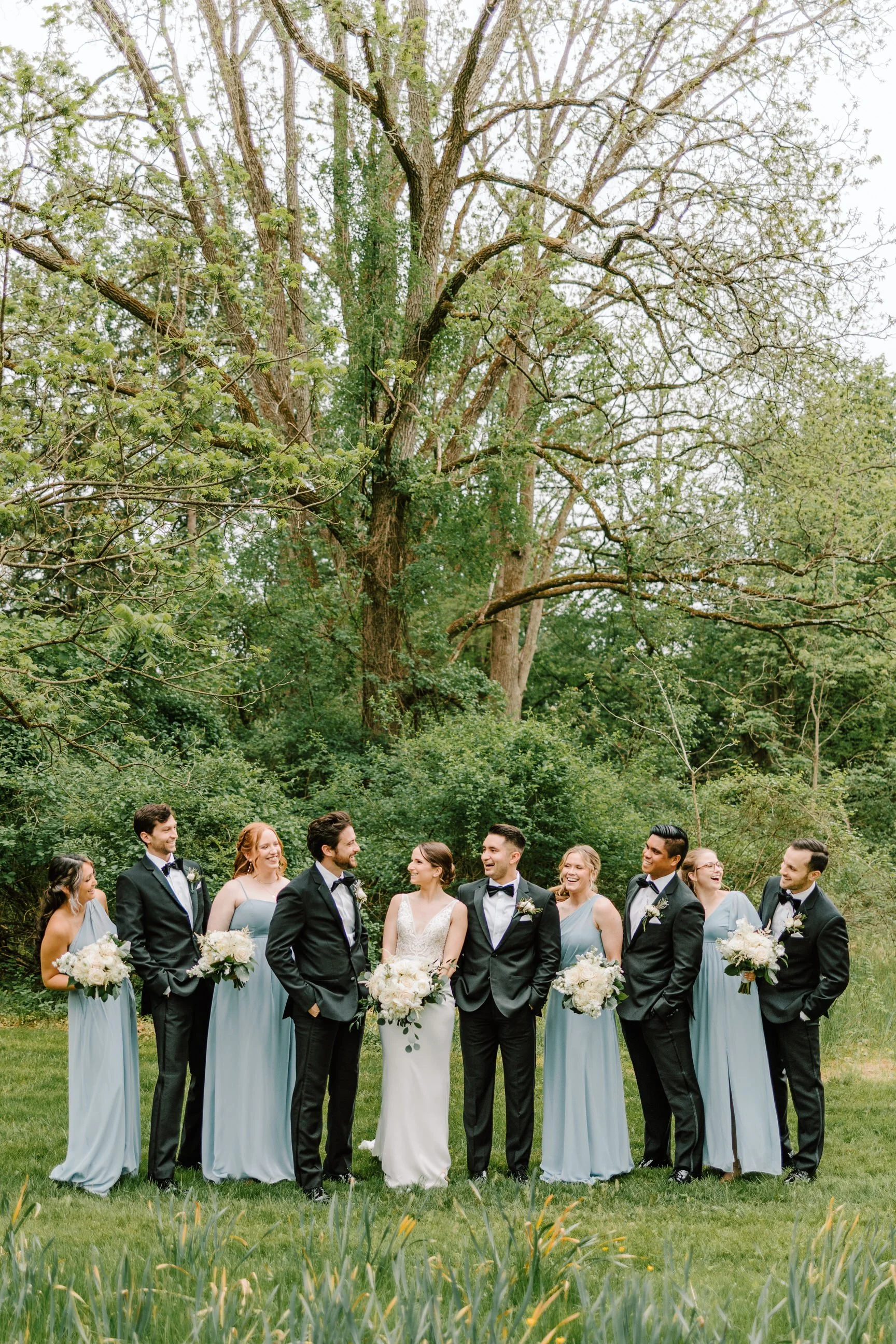 A wedding party standing outdoors on grass with trees in the background, including a bride in a white gown, bridesmaids in light blue dresses, and groomsmen in black suits with bow ties, holding bouquets and smiling.