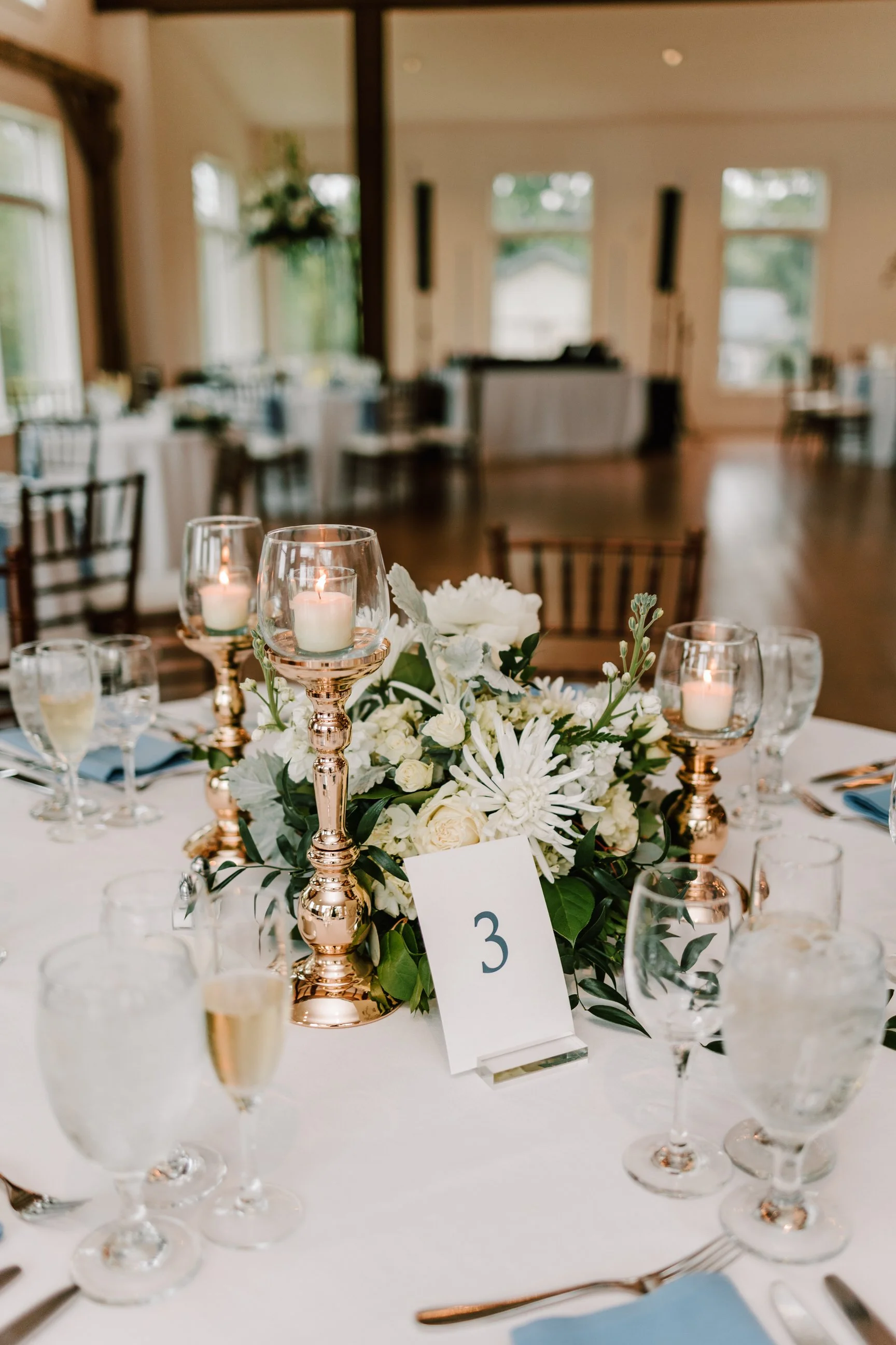 Elegant round table at a wedding reception with a floral centerpiece of white flowers and green leaves, surrounded by rose gold candle holders with white candles, wine glasses, champagne glasses, and table number '3' in an upscale banquet hall with l