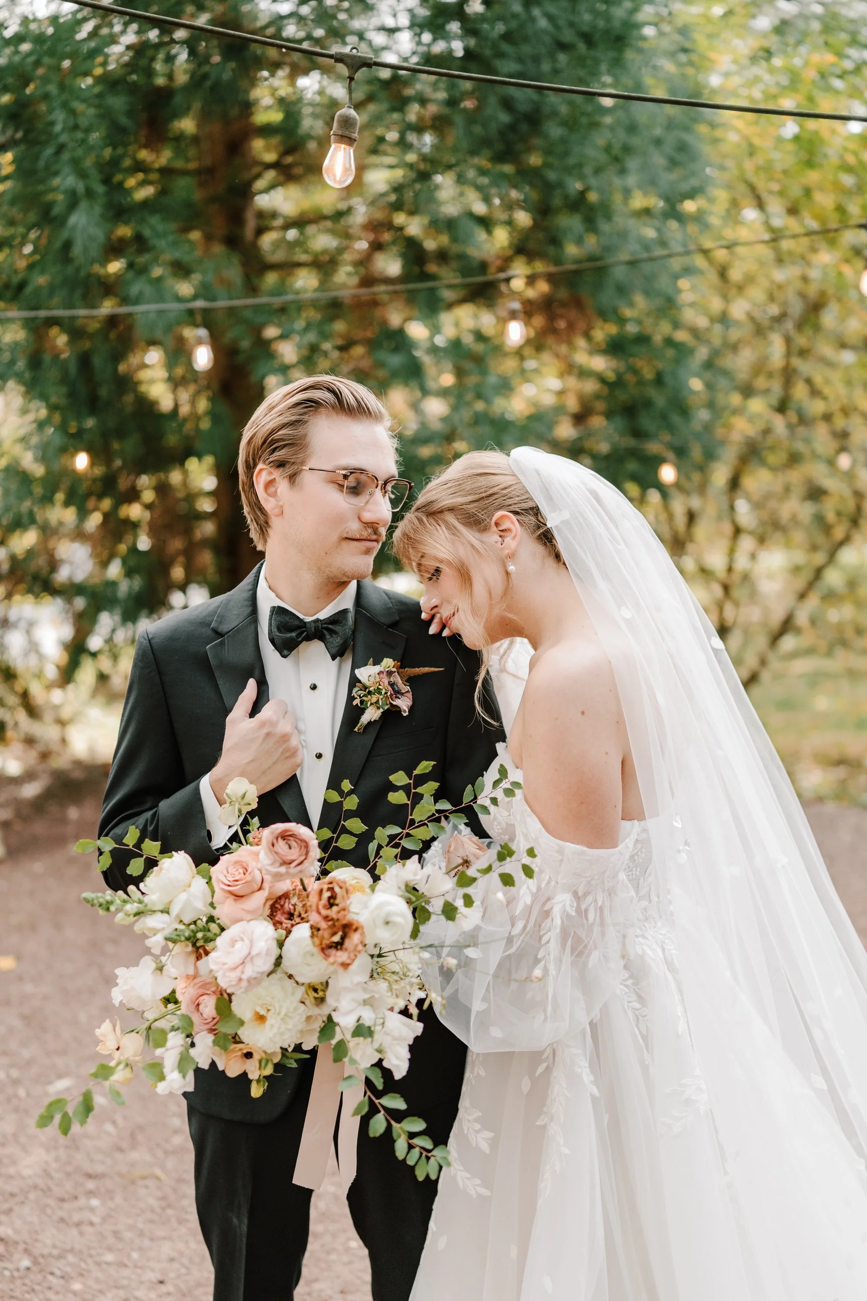 A bride and groom sharing an intimate moment outdoors, with the groom in a tuxedo holding a bouquet of flowers, and the bride leaning her head on his shoulder, wearing a wedding dress and veil, surrounded by trees and string lights. Holly Hedge
