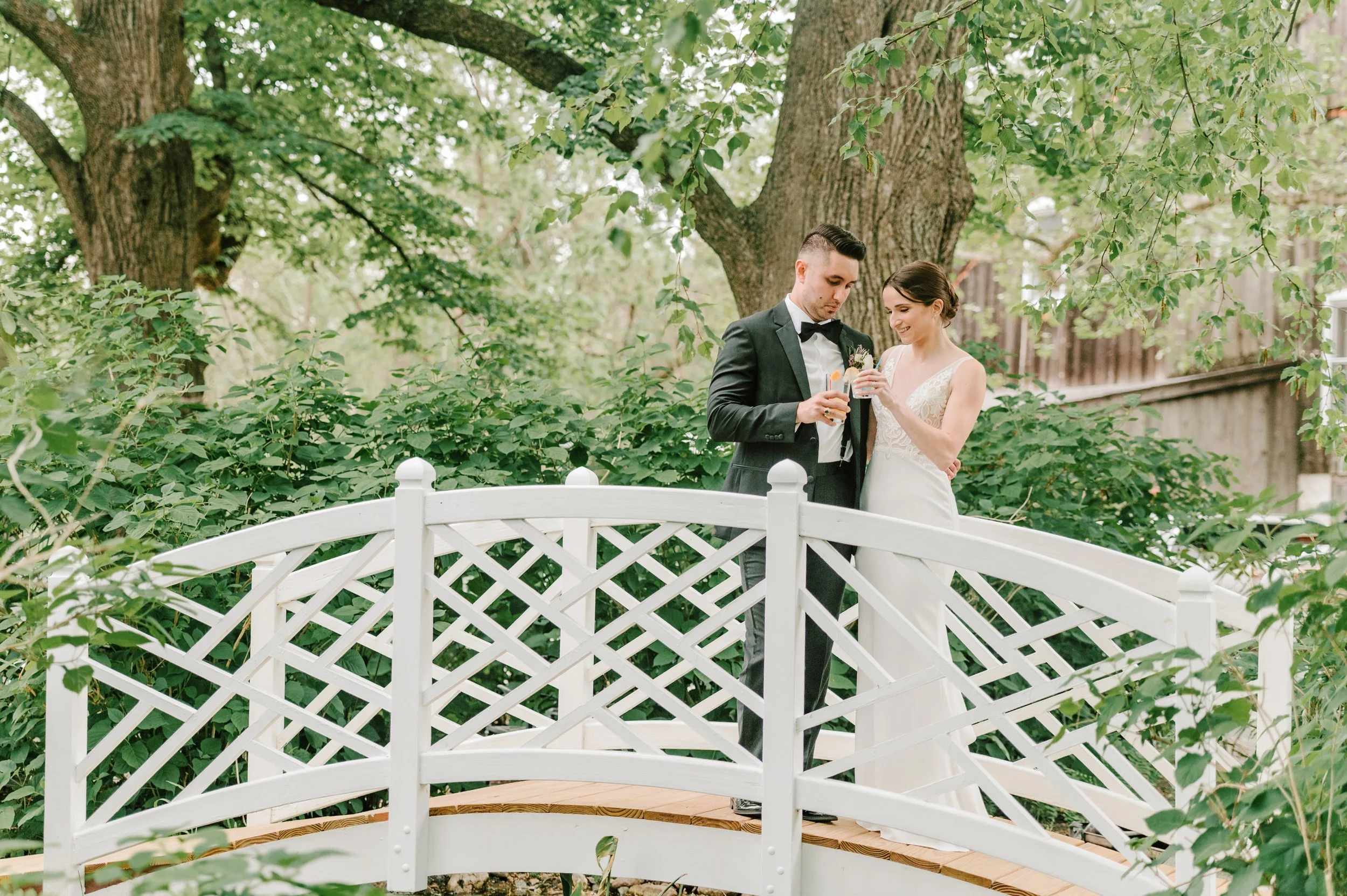 A newlywed couple on a white decorative bridge in a lush green garden, holding glasses and smiling at each other.