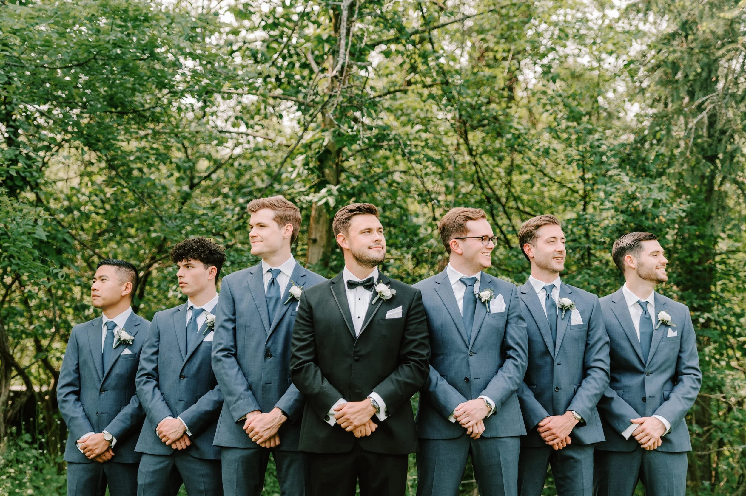 Group of seven men in suits standing outdoors on green foliage background, with the groom in a black tuxedo in the center, all smiling and looking in different directions.
