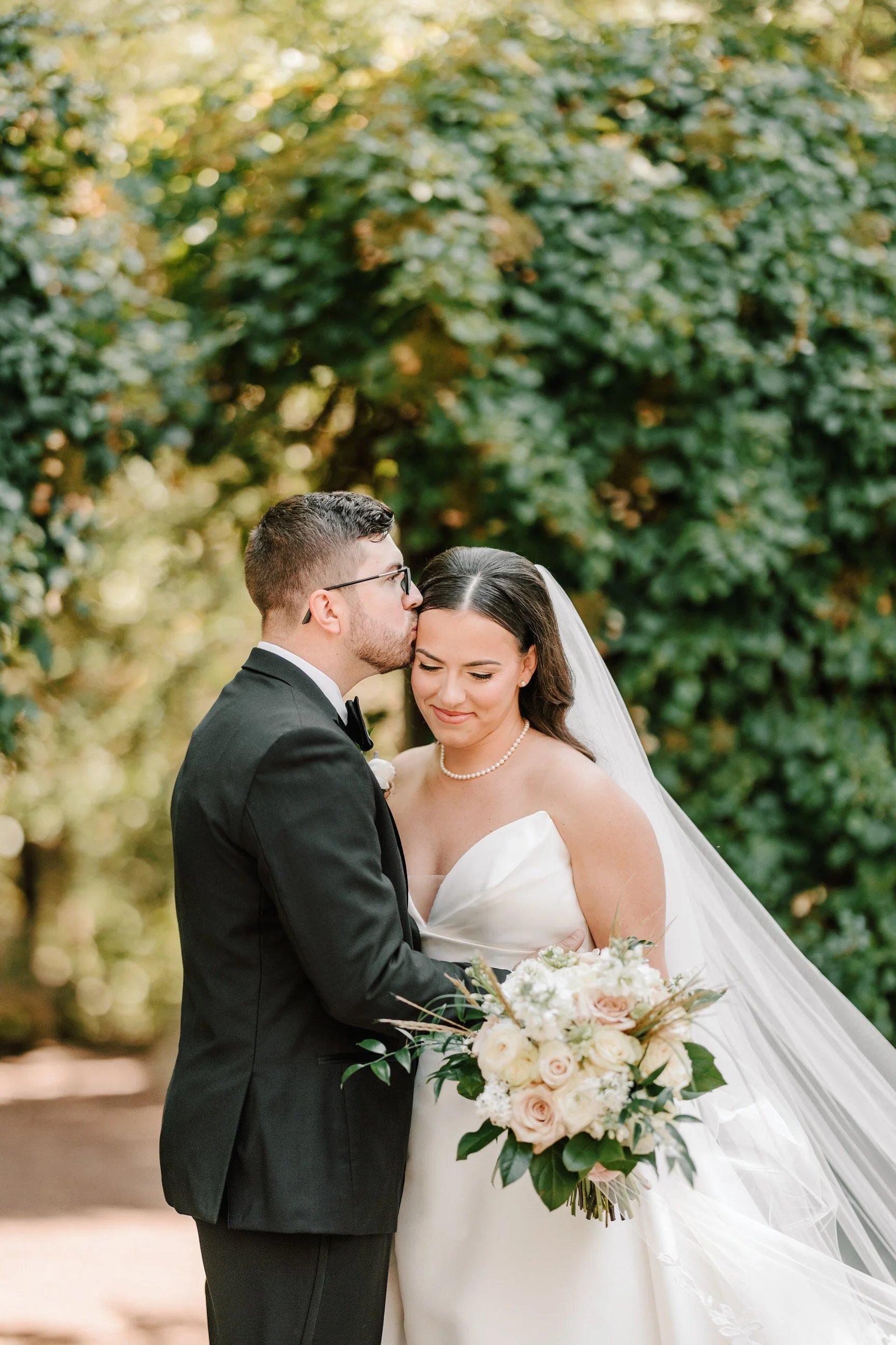 A groom kissing a bride on the forehead during a wedding, with the bride holding a bouquet of white and blush roses, wearing a strapless wedding gown and pearl necklace, outdoors with green foliage in the background.