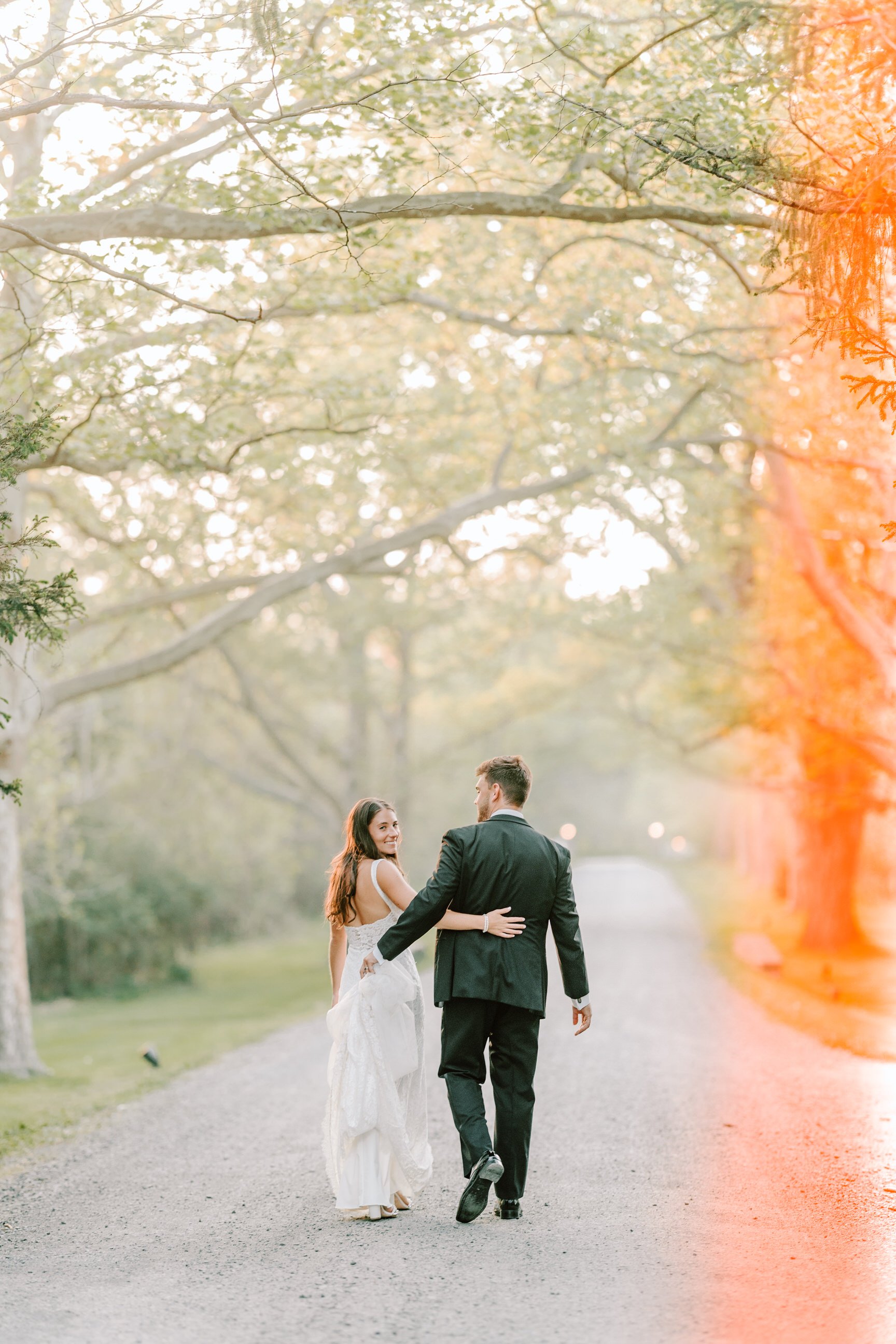 A bride and groom walking together on a gravel path in a park-like setting, holding each other with trees and soft sunlight in the background.