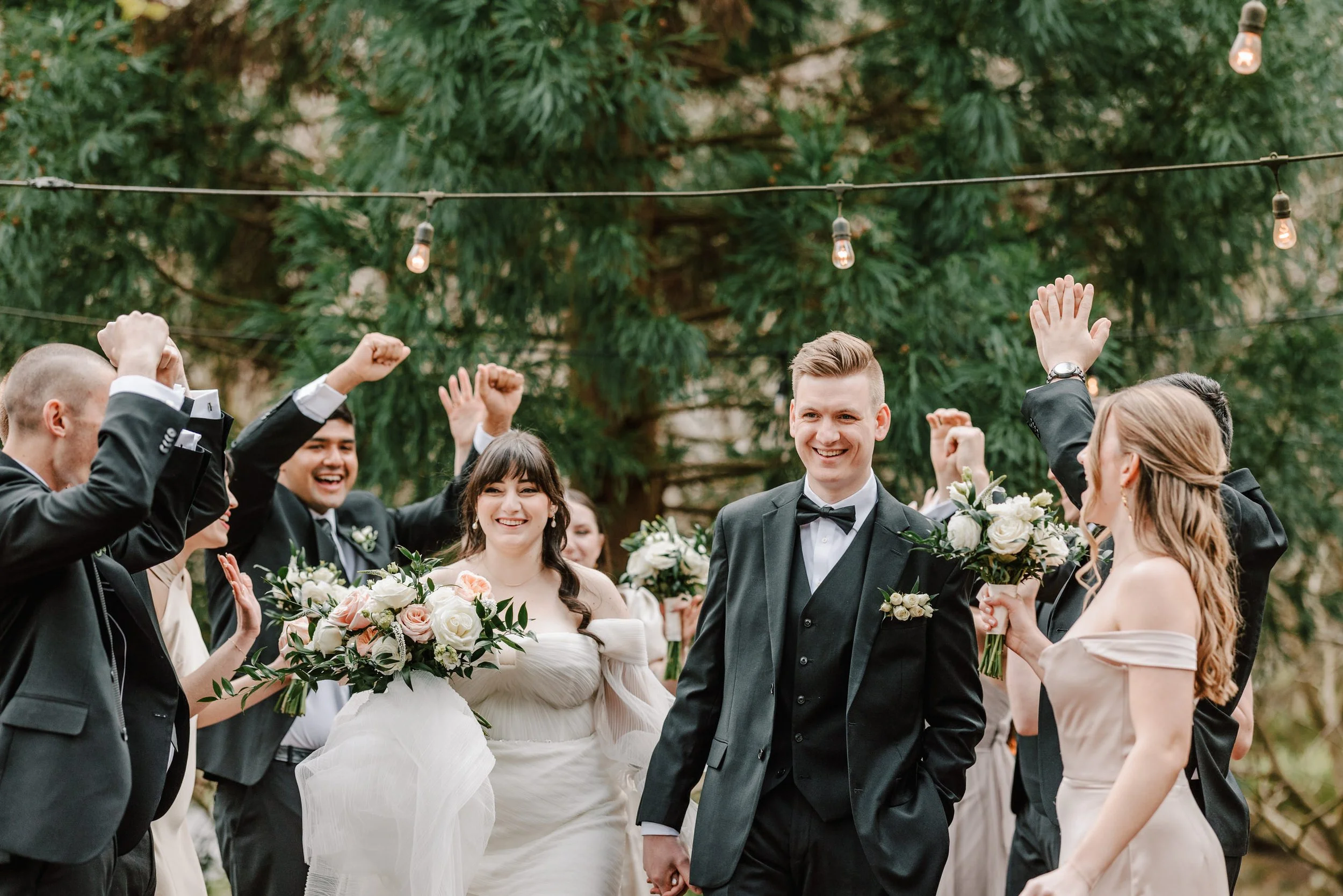 A bride and groom celebrating with their wedding party outdoors under string lights. The couple is holding hands and smiling, while the bridesmaids and groomsmen cheer around them, holding bouquets of white and peach flowers.