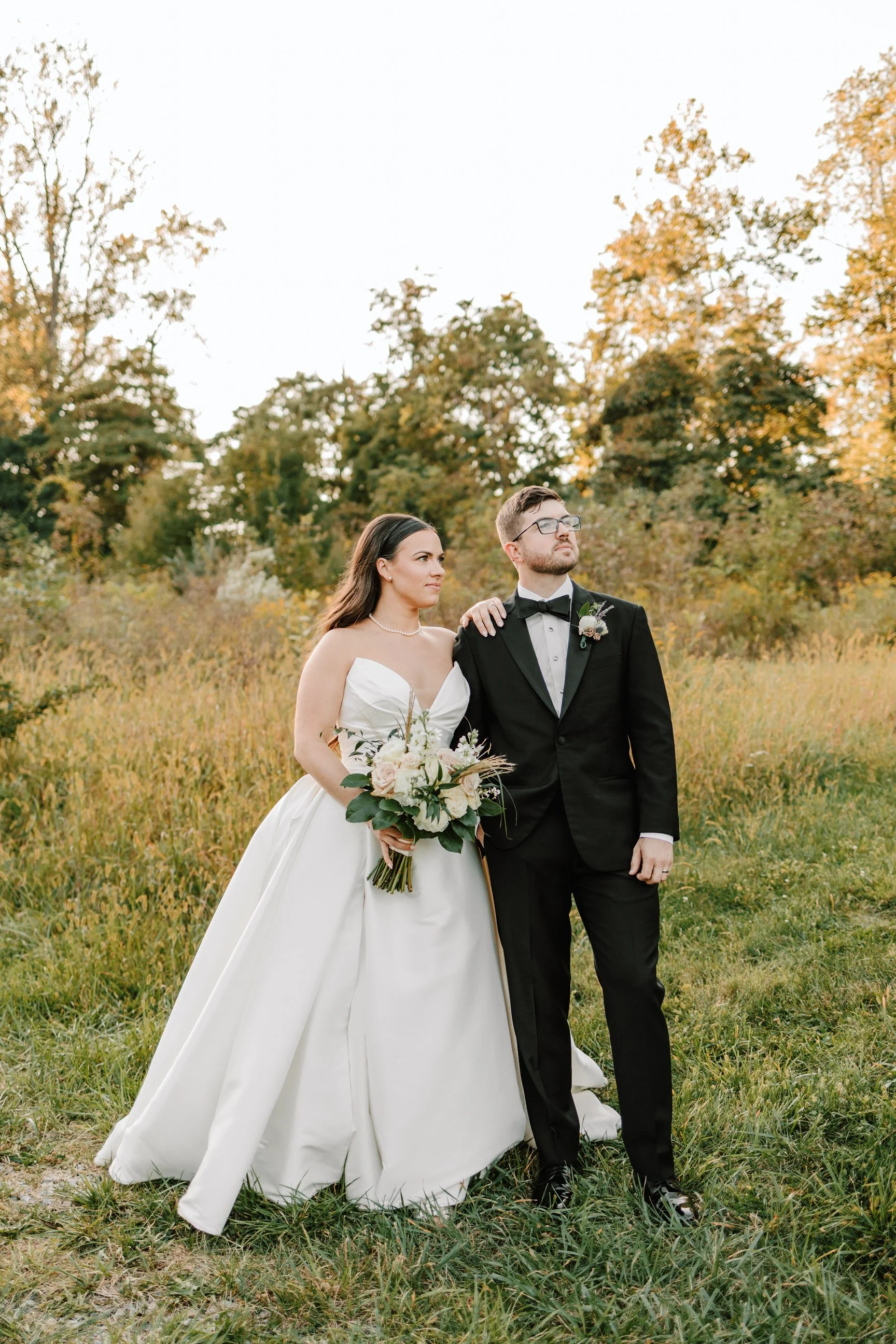 Bride and groom standing outdoors in a grassy area with autumn trees in the background. The bride wears a white wedding gown and holds a bouquet, while the groom wears a black tuxedo with a bow tie.