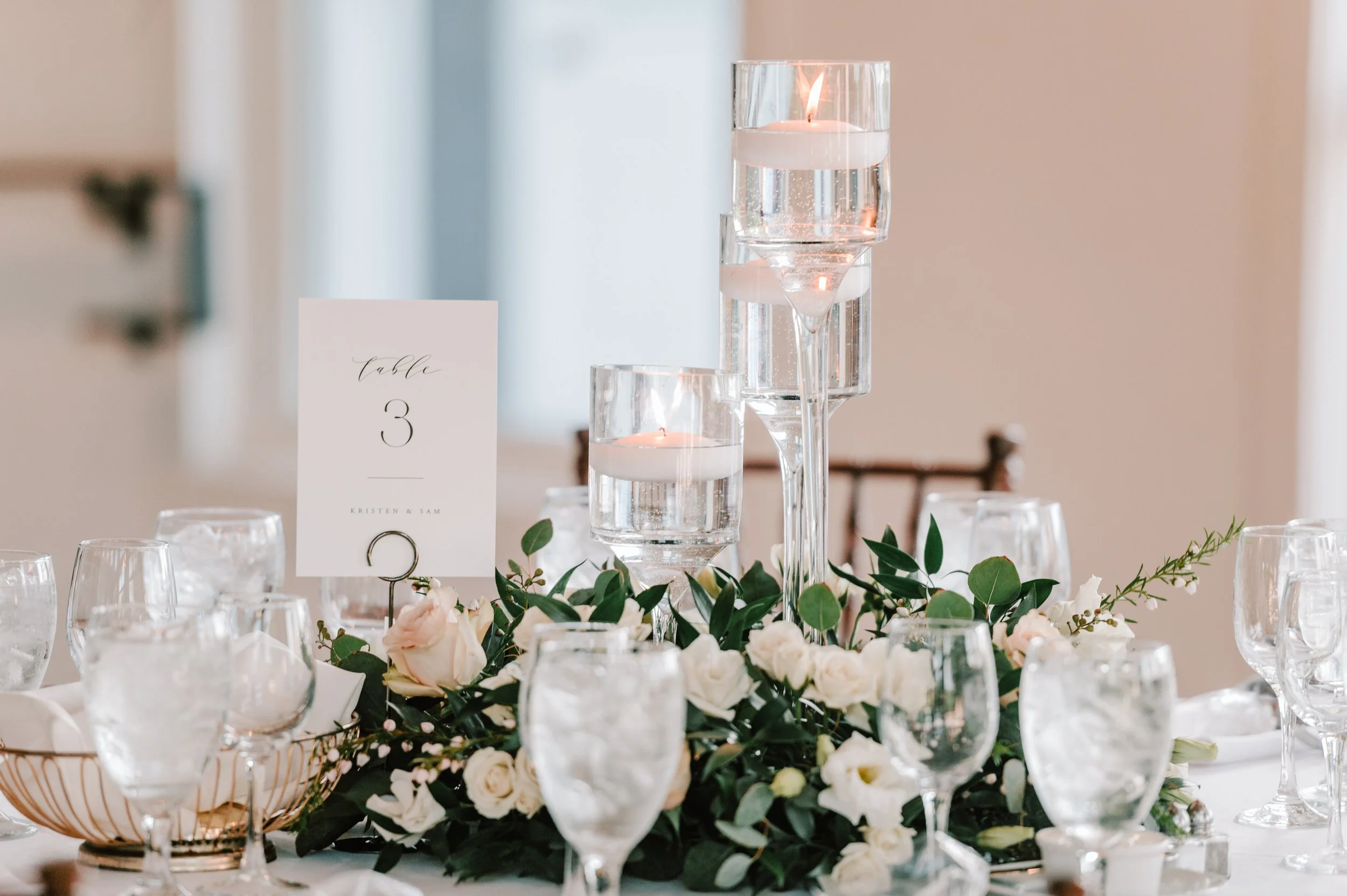 Elegant wedding table centerpiece with candles, greenery, and white flowers, with a table number card and glassware.