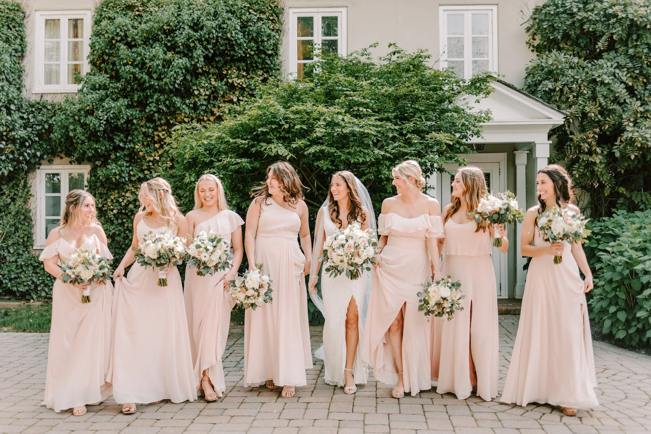 A bride and her eight bridesmaids walking on a brick pathway in front of a house covered in ivy. They are smiling and holding bouquets of flowers. The bride is in the center, wearing a wedding gown, and the bridesmaids are in matching blush pink dres