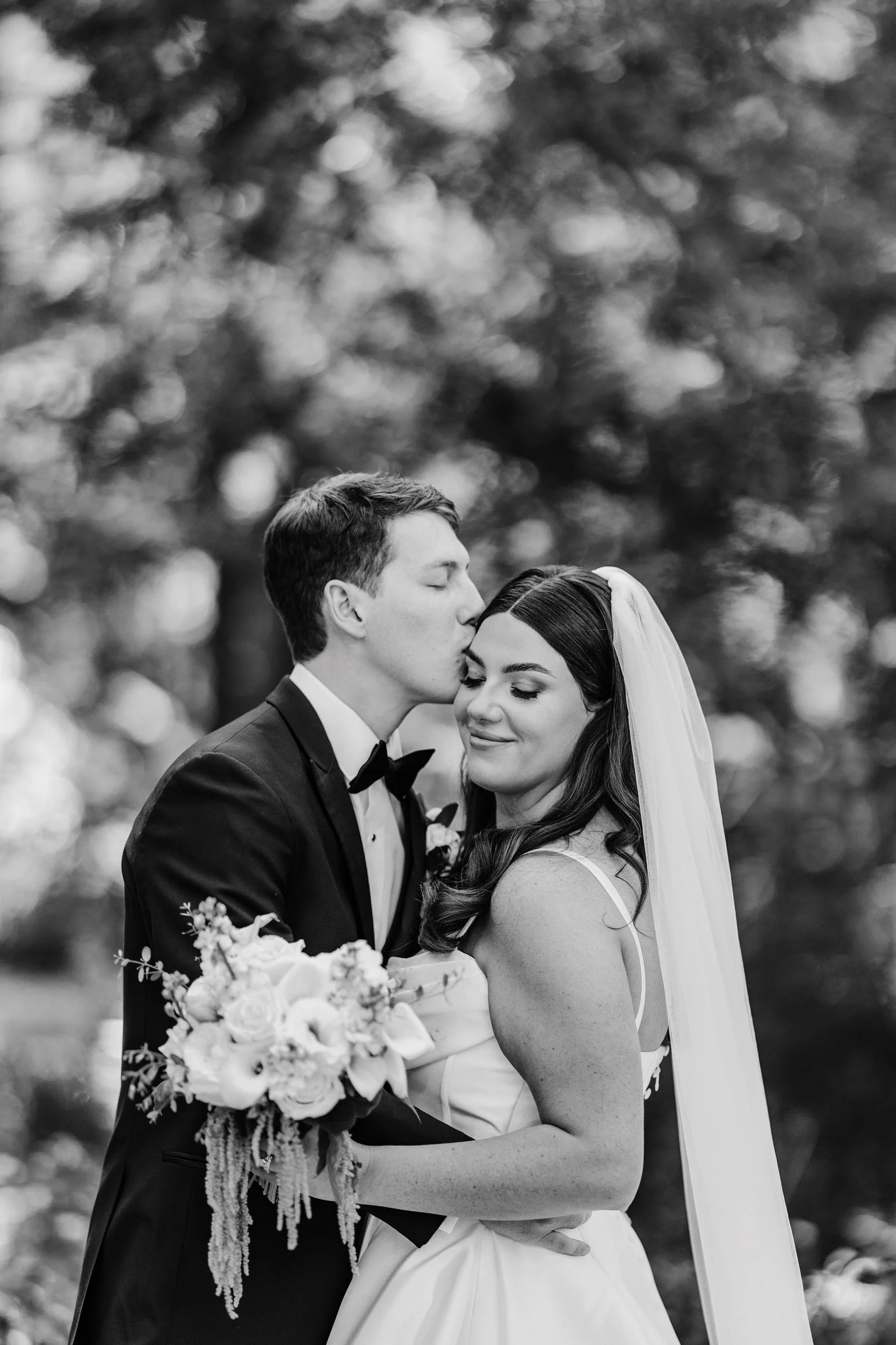 Black and white photograph of a bride and groom embracing outdoors. The groom is kissing the bride's forehead while she smiles with her eyes closed, holding a bouquet of flowers. The bride has dark hair, and the groom is wearing a tuxedo with a bow t