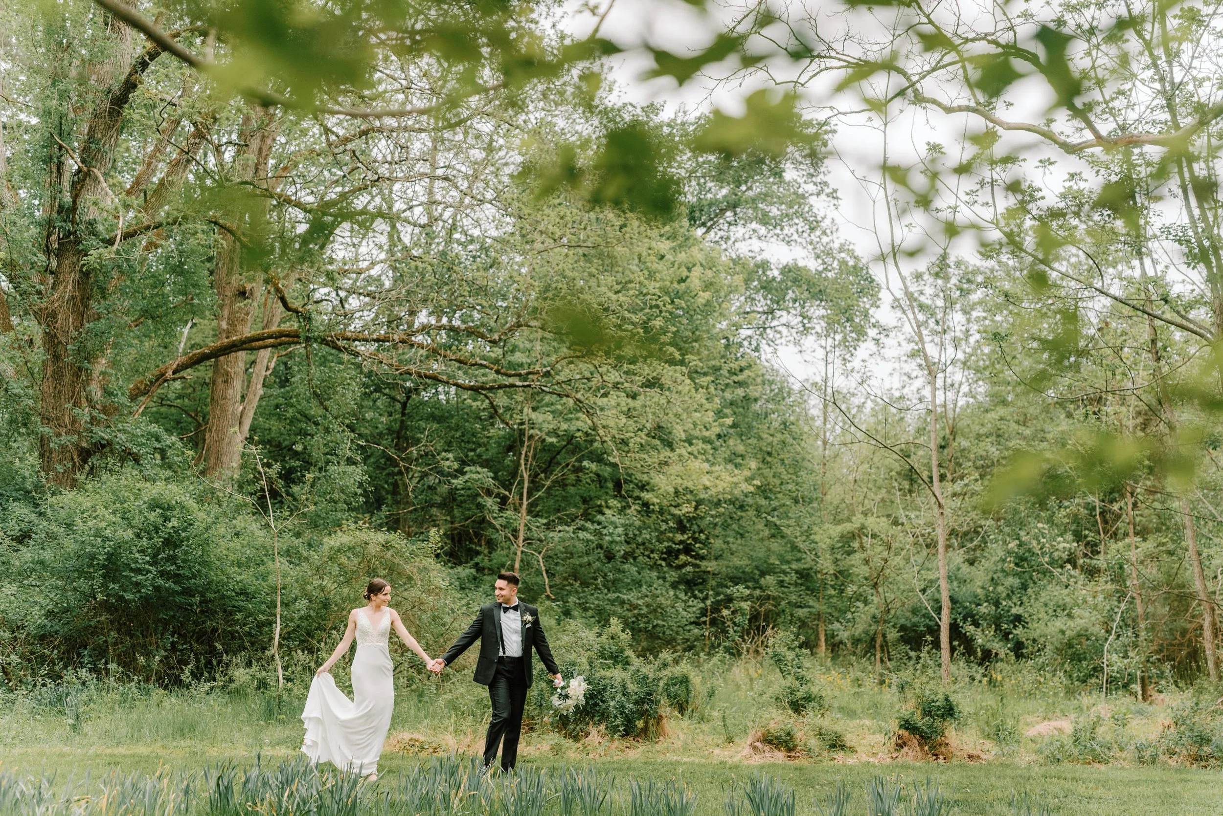 A bride and groom walking hand in hand through a lush, green outdoor setting with trees and grass, dressed in wedding attire.