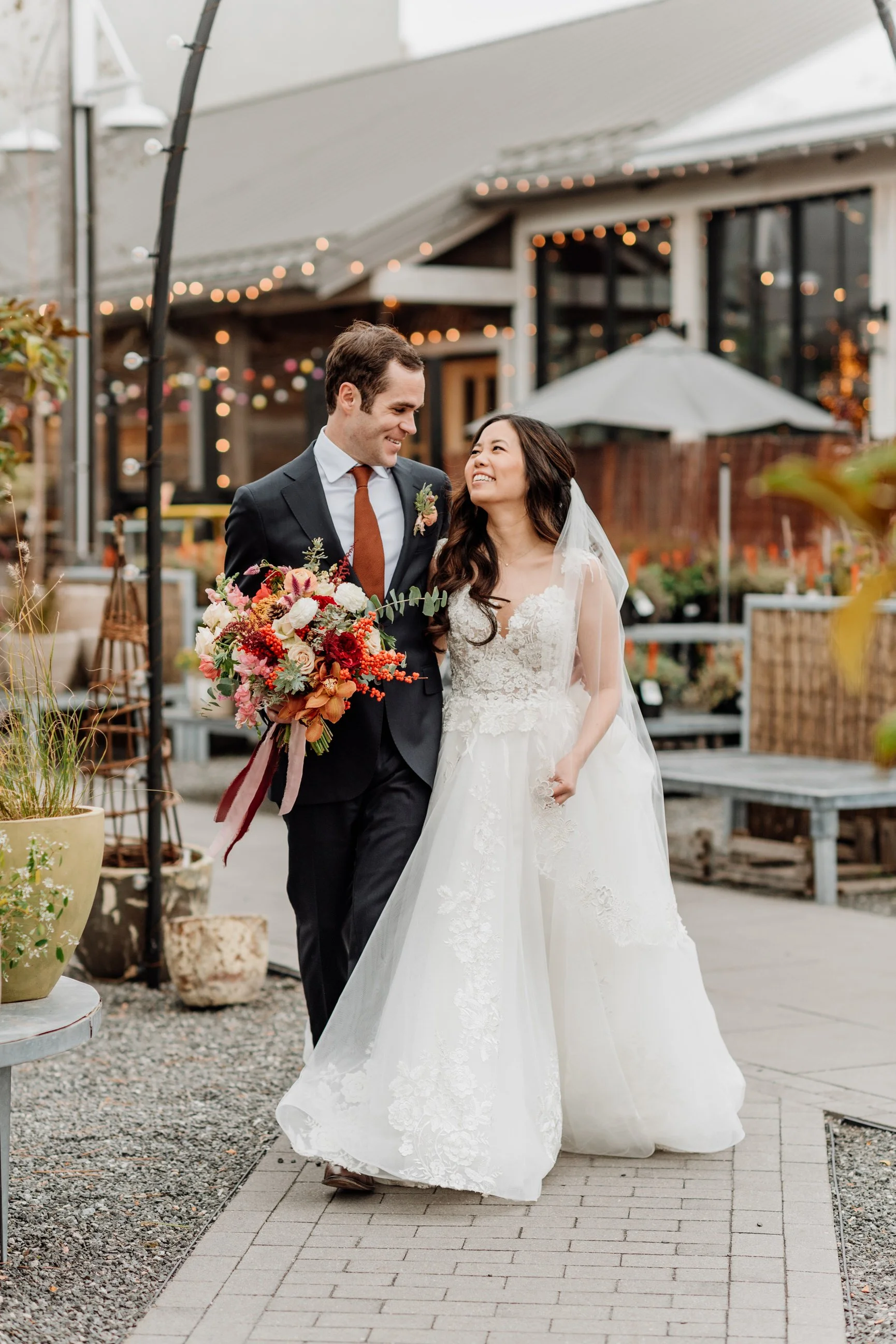 A newlywed couple walking outdoors at dusk, with the groom in a black suit and tie and the bride in a white lace wedding gown and veil, holding a large bouquet of red, pink, and orange flowers, smiling at each other.