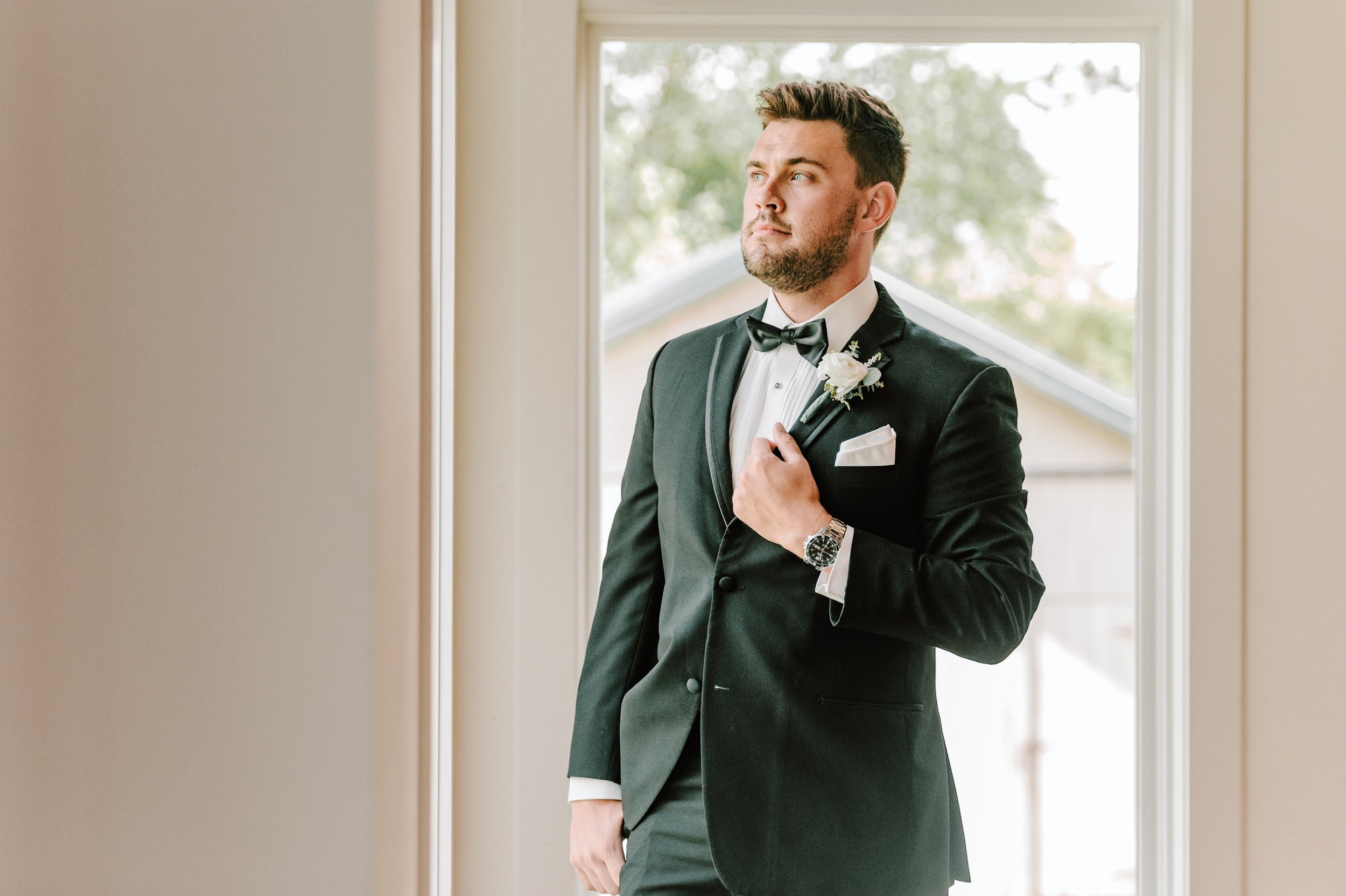 A man dressed in a black tuxedo with a bow tie and white shirt, standing inside near a window, holding a flower boutonniere and looking off to the side.