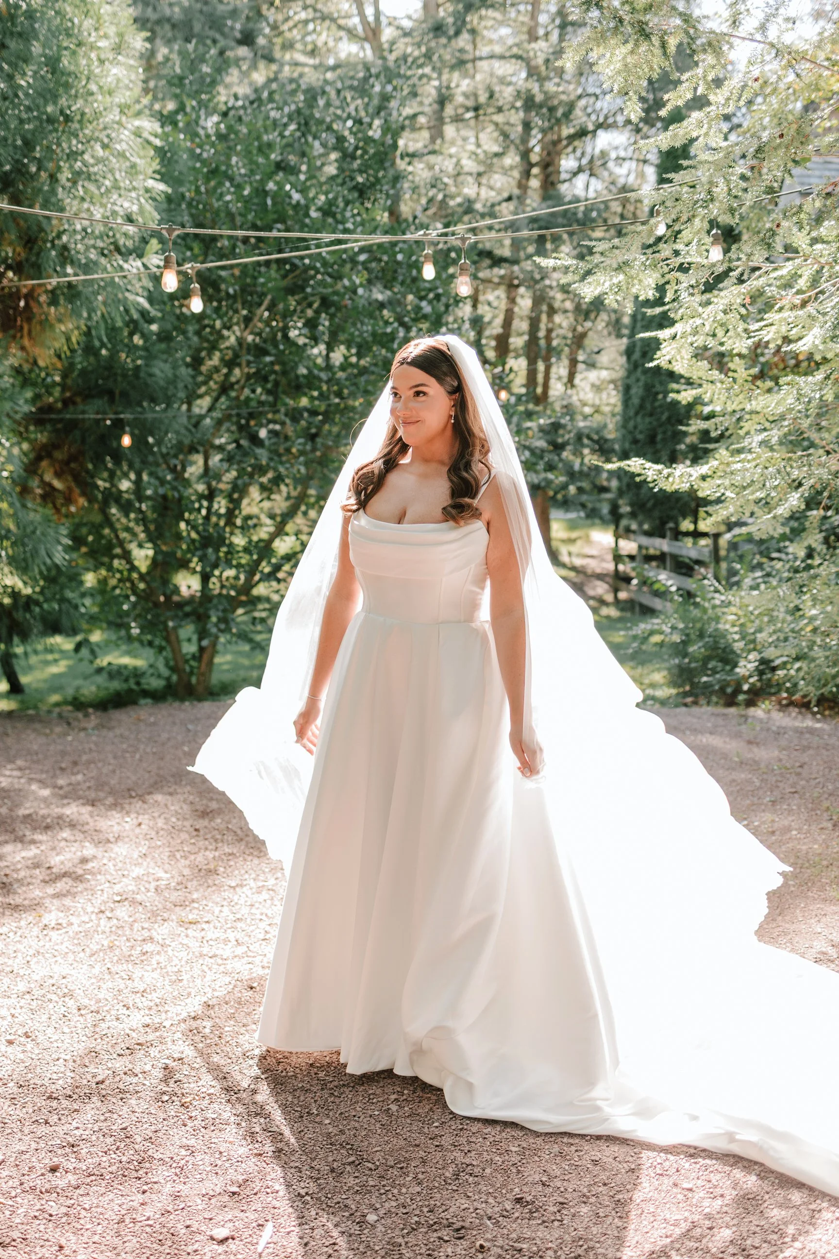 A bride in a white wedding gown and veil stands outdoors in a wooded area, illuminated by sunlight and decorative string lights.