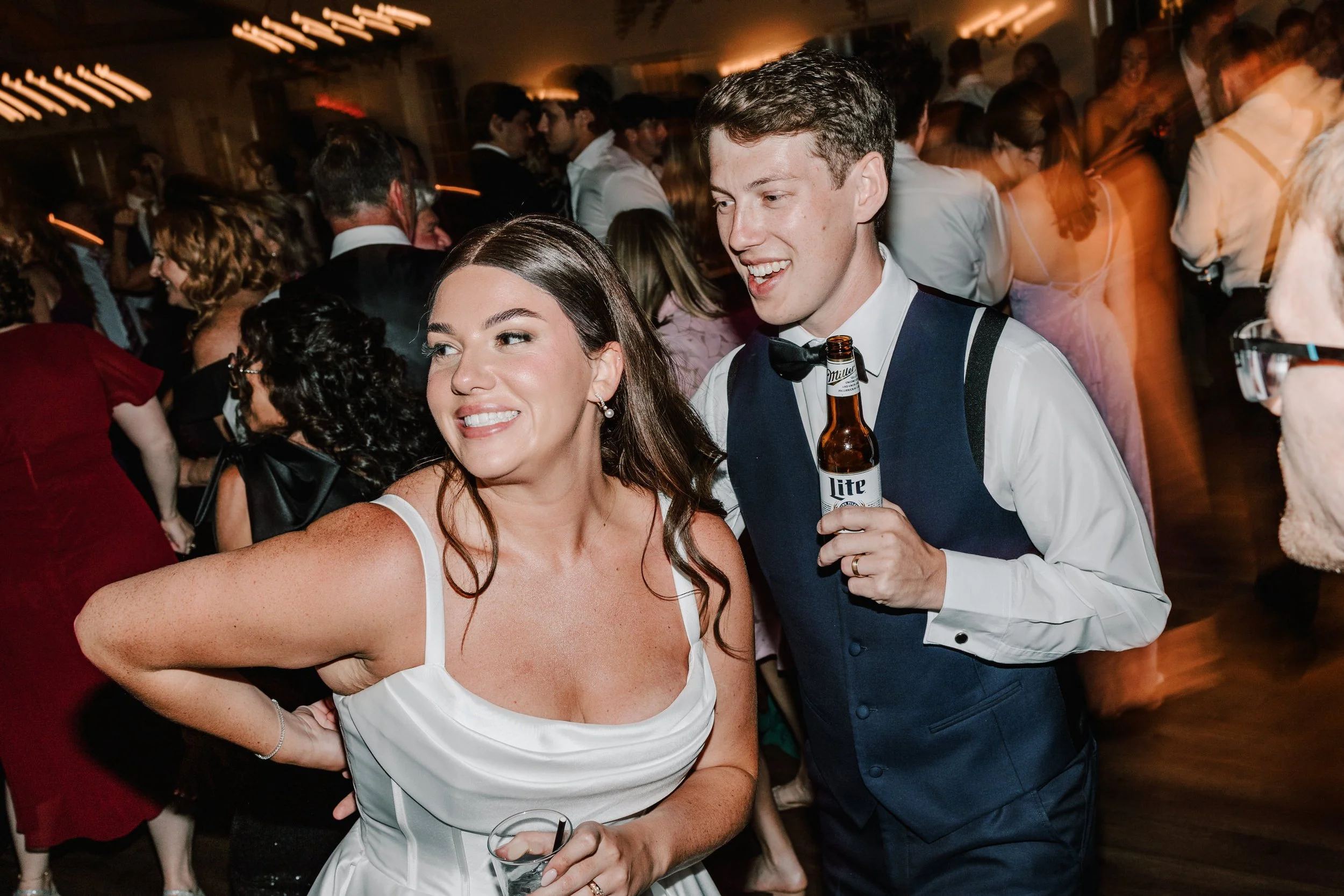 A woman in a white dress smiling and holding a drink, standing next to a man in a tuxedo with a bow tie, who is holding a beer, at a lively party or wedding reception.