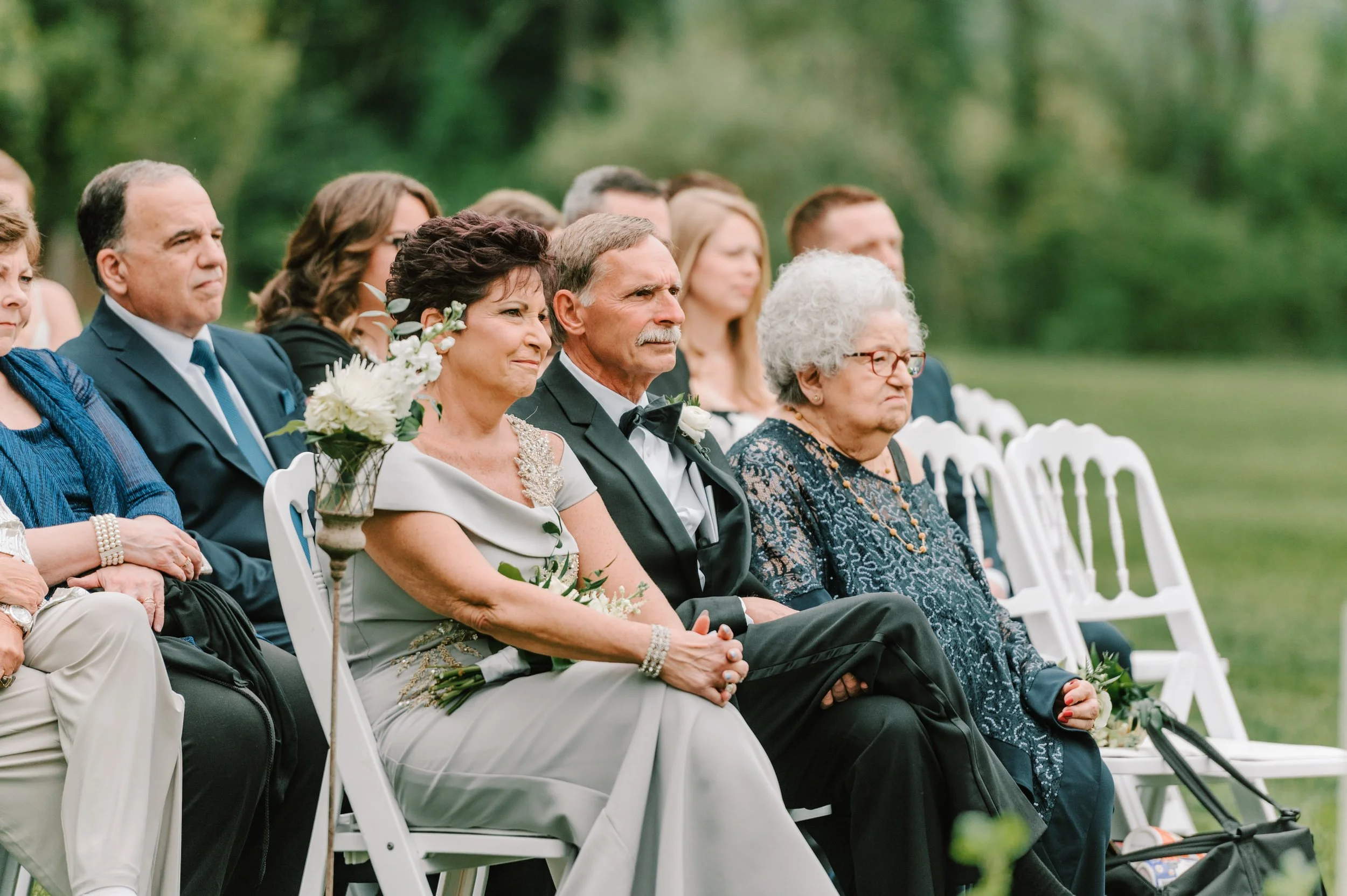 Group of older people sitting outdoors at a wedding ceremony, dressed in formal attire, with a lush green background.