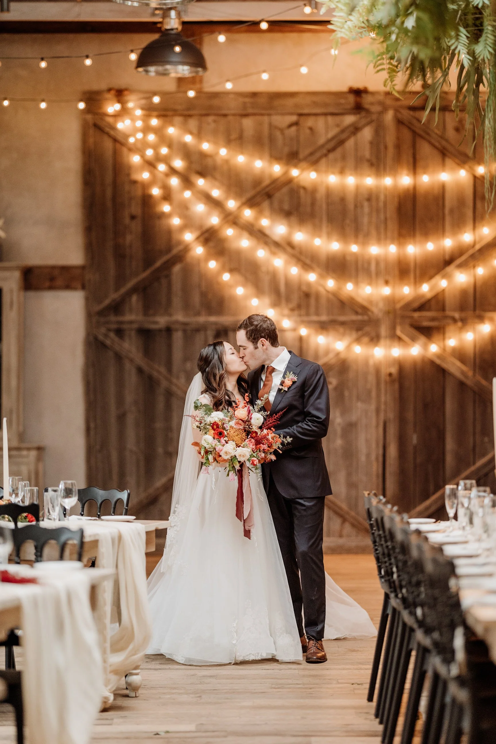 A bride and groom share a kiss during their wedding reception, with string lights and a wooden barn door in the background.