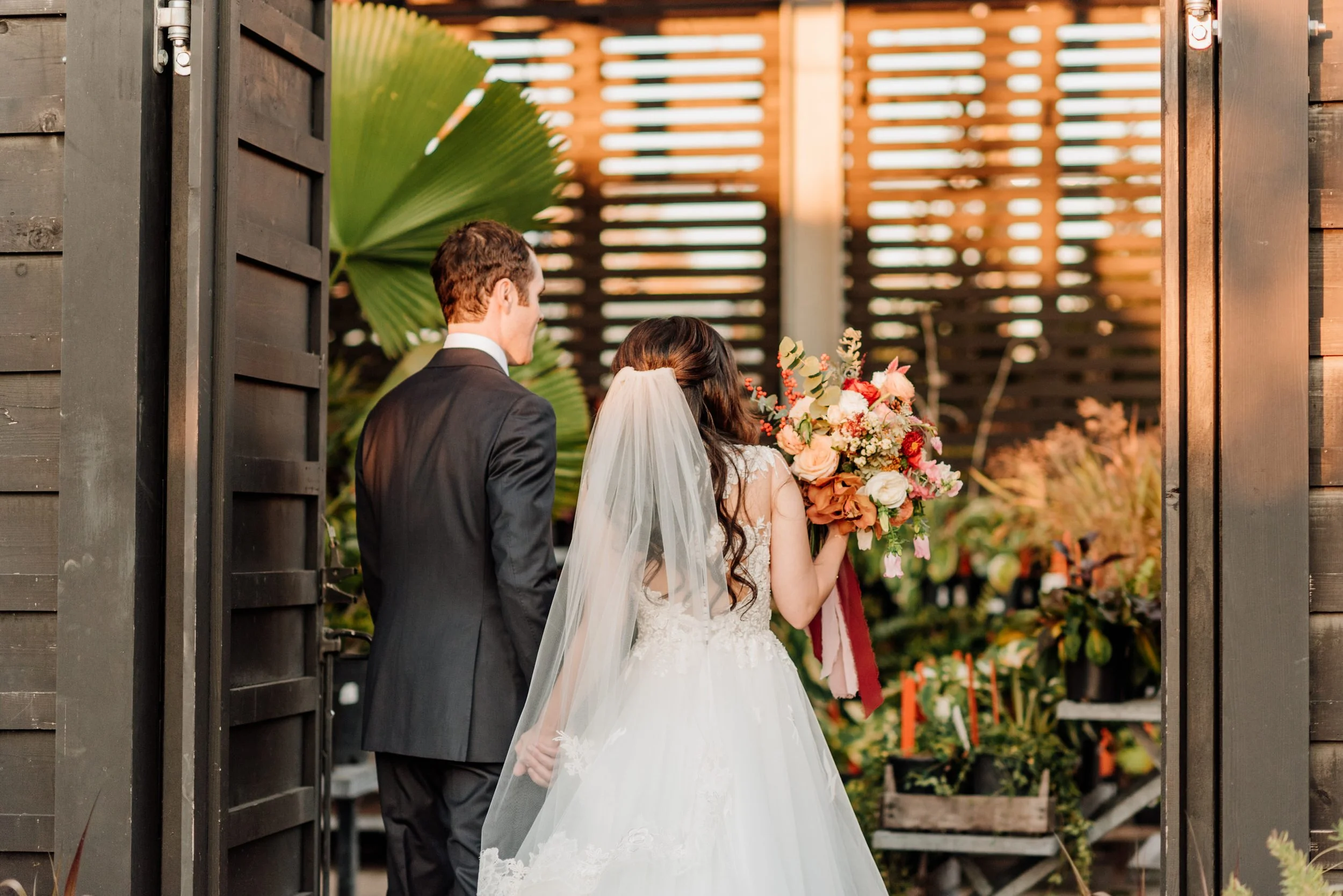 A bride in a white wedding dress holding a bouquet of flowers standing next to a groom in a black suit, viewed from behind, in an outdoor setting with wooden walls and greenery.