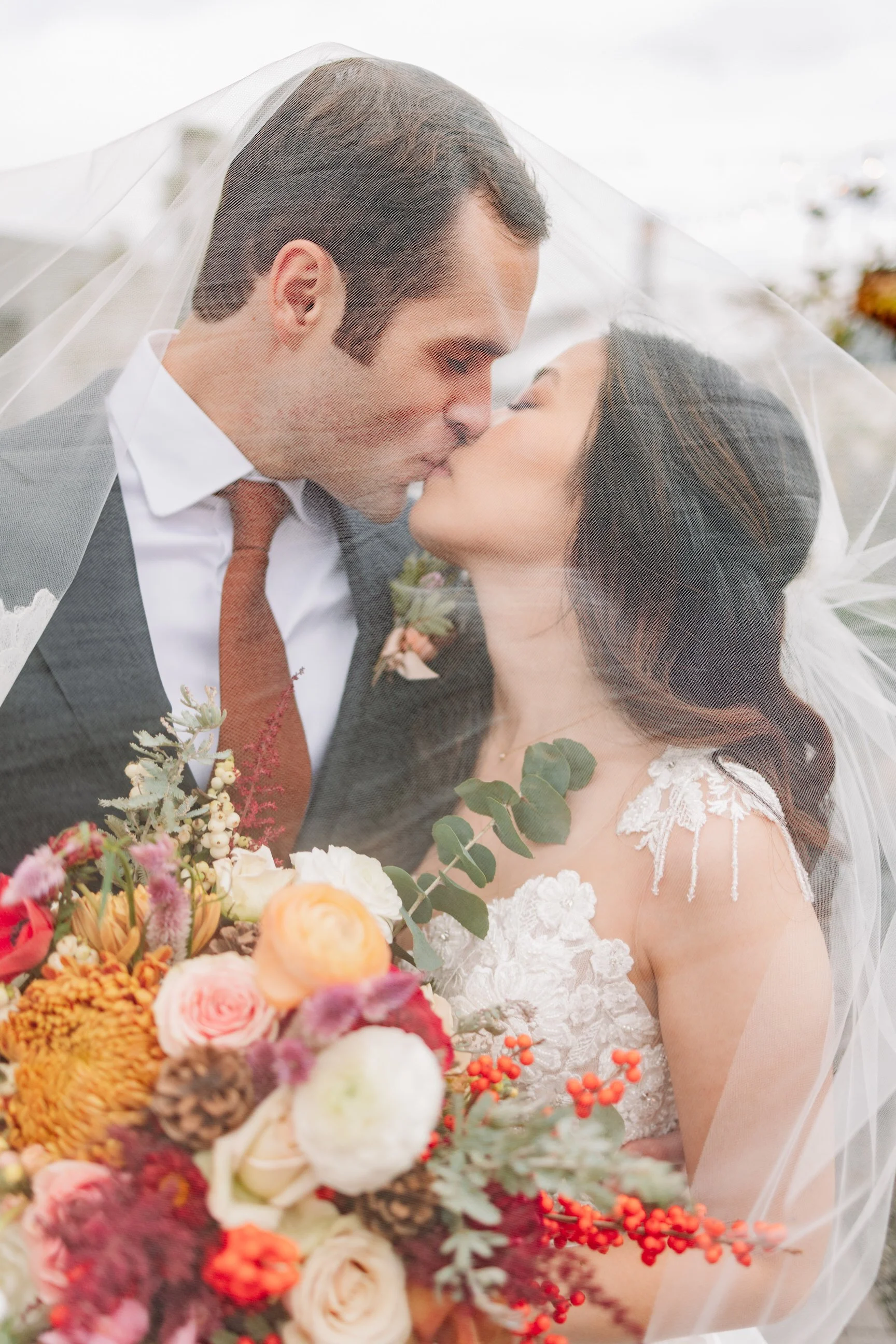 A bride and groom kiss under a sheer veil, holding a bouquet of colorful flowers, with an outdoor background.