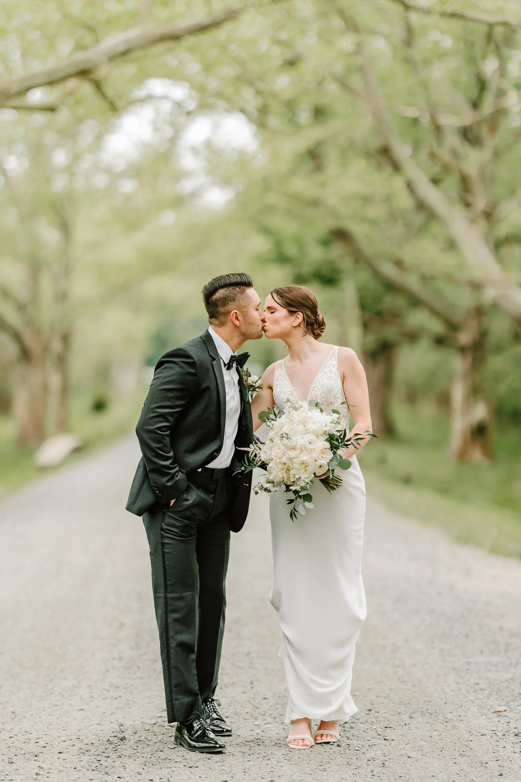 A bride and groom sharing a kiss outdoors on a tree-lined pathway, with the bride holding a bouquet of white flowers and greenery.