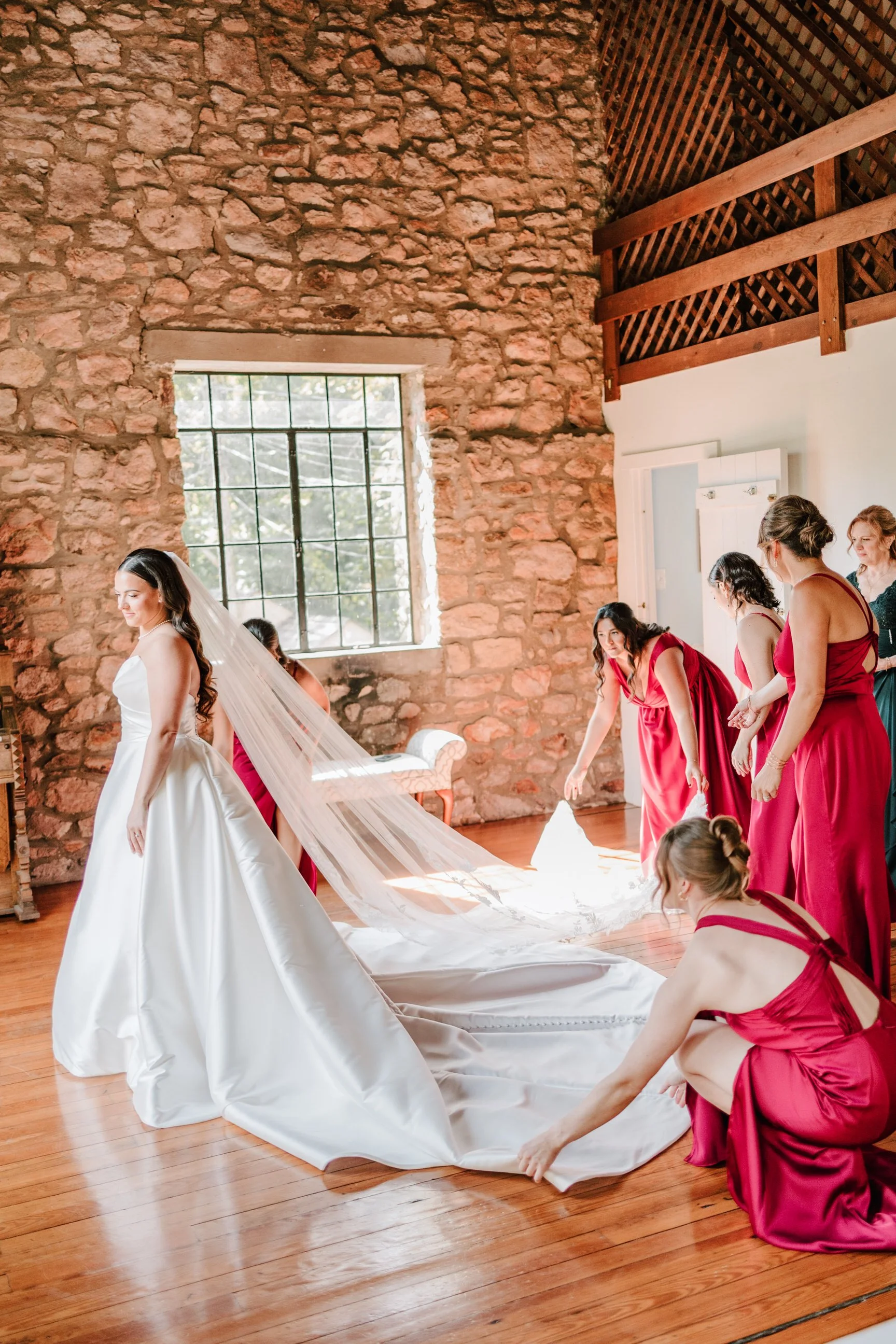 A bride in a white wedding gown and veil stands in a rustic room with exposed stone walls, while her bridesmaids in red dresses help arrange her long train and veil.