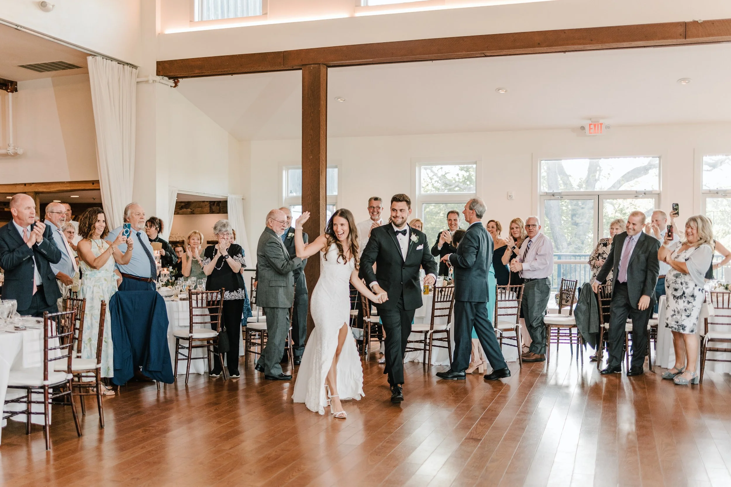 A bride and groom entering a wedding reception hall, walking hand in hand while guests applaud and take pictures, with a bright and airy venue featuring wooden floors and large windows.