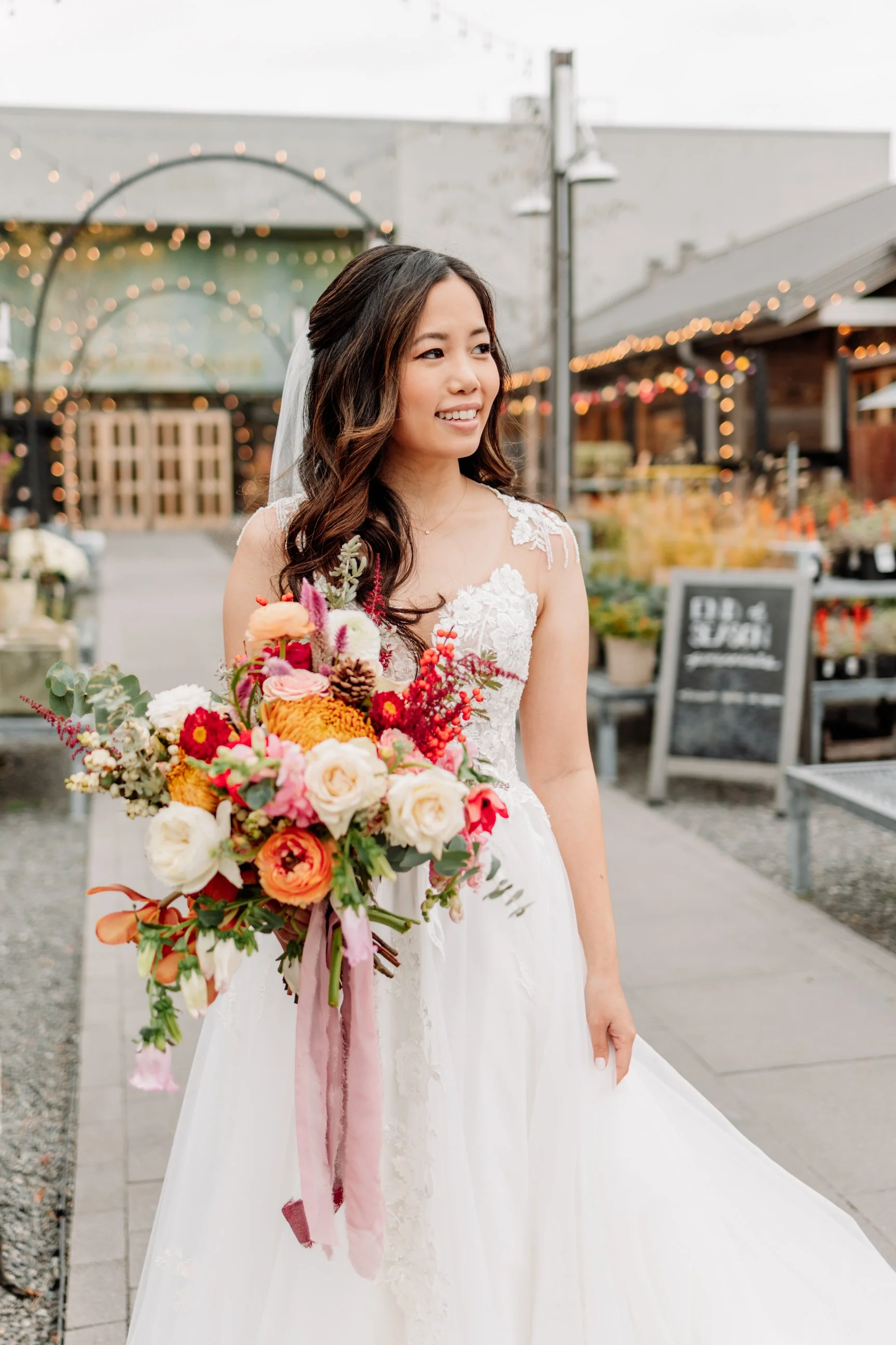 A bride in a white wedding dress holding a colorful bouquet of flowers outside at a venue decorated with string lights.