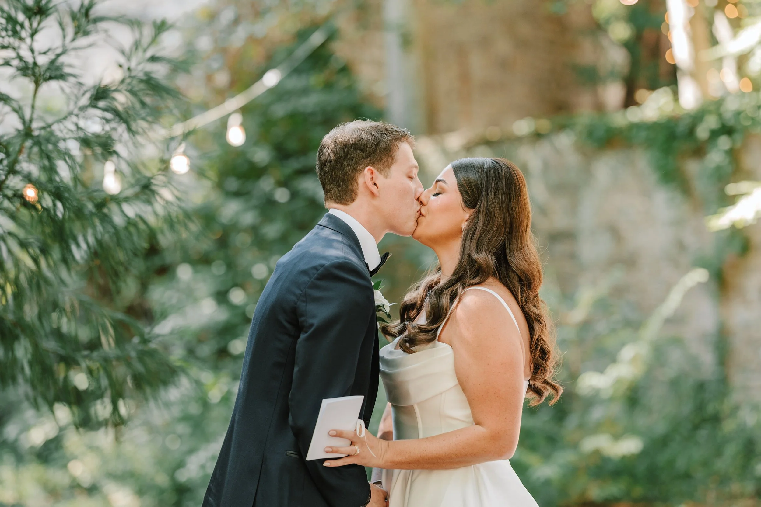 A bride and groom kiss outdoors during their wedding ceremony, surrounded by greenery and string lights.