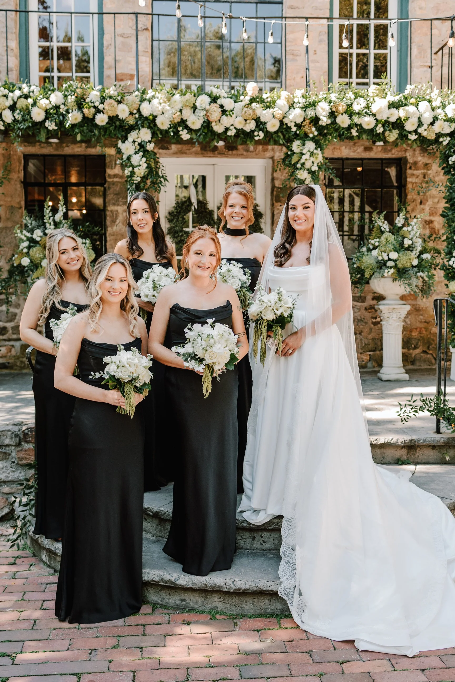 A bride and her bridesmaids pose for a wedding photo outdoors in front of a decorated building with floral arrangements and string lights.