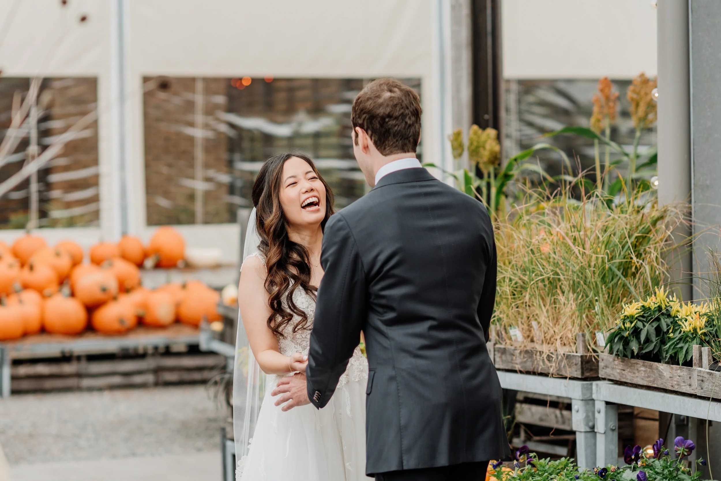 A bride and groom share a joyful moment inside a greenhouse or plant shop. The bride is wearing a white wedding dress and smiling while looking at the groom, who is in a black suit. Pumpkins and plants are visible in the background.