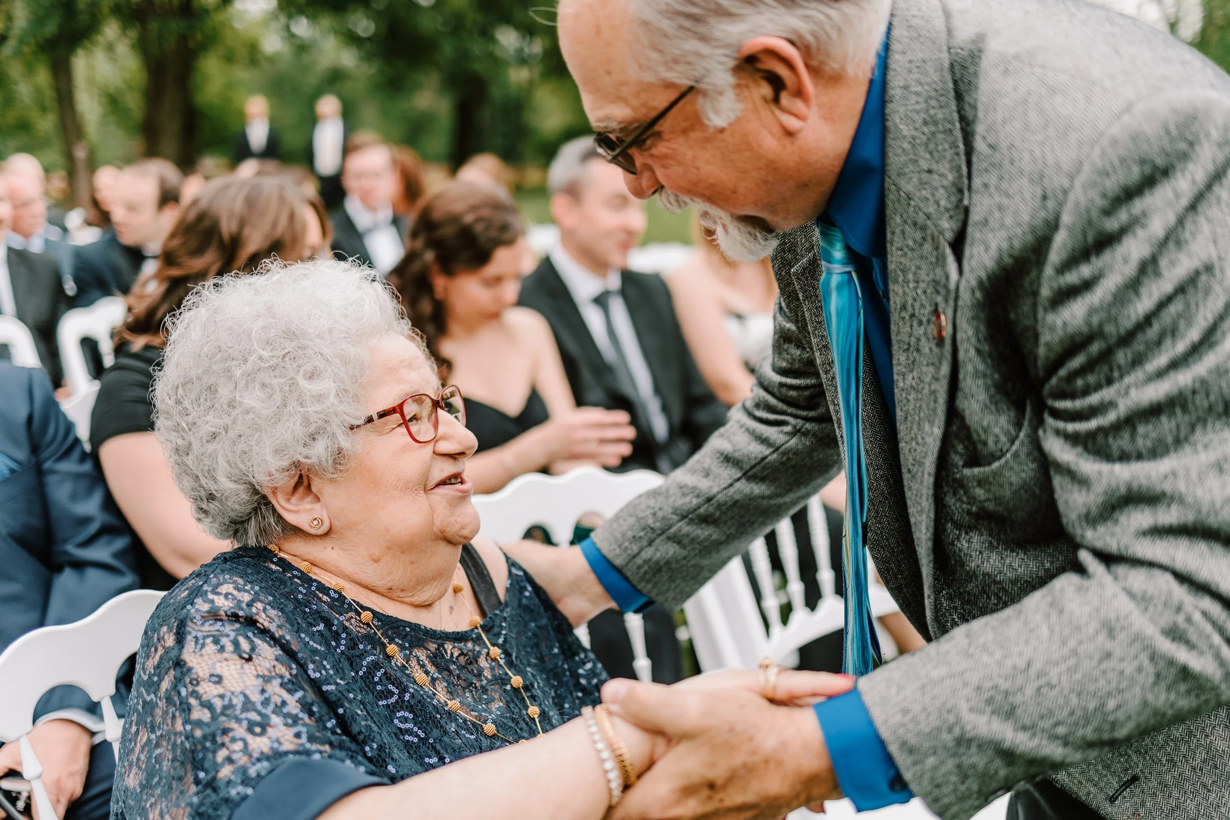 An elderly woman with curly gray hair and glasses smiling as she holds hands with a man in a gray suit and blue shirt at an outdoor wedding ceremony. People in formal attire are seated in the background.