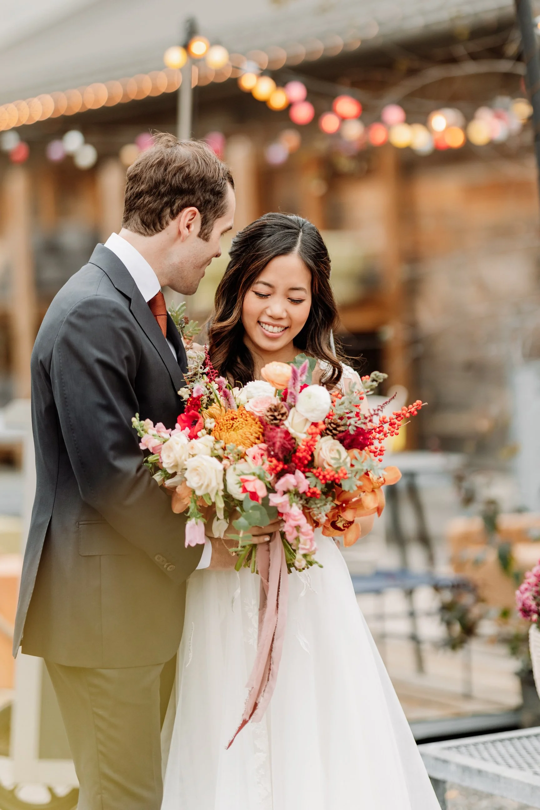 A bride holding a large bouquet of colorful flowers, smiling as she looks down, standing next to a groom in a gray suit holding the bouquet in an outdoor setting with string lights and a rustic backdrop.