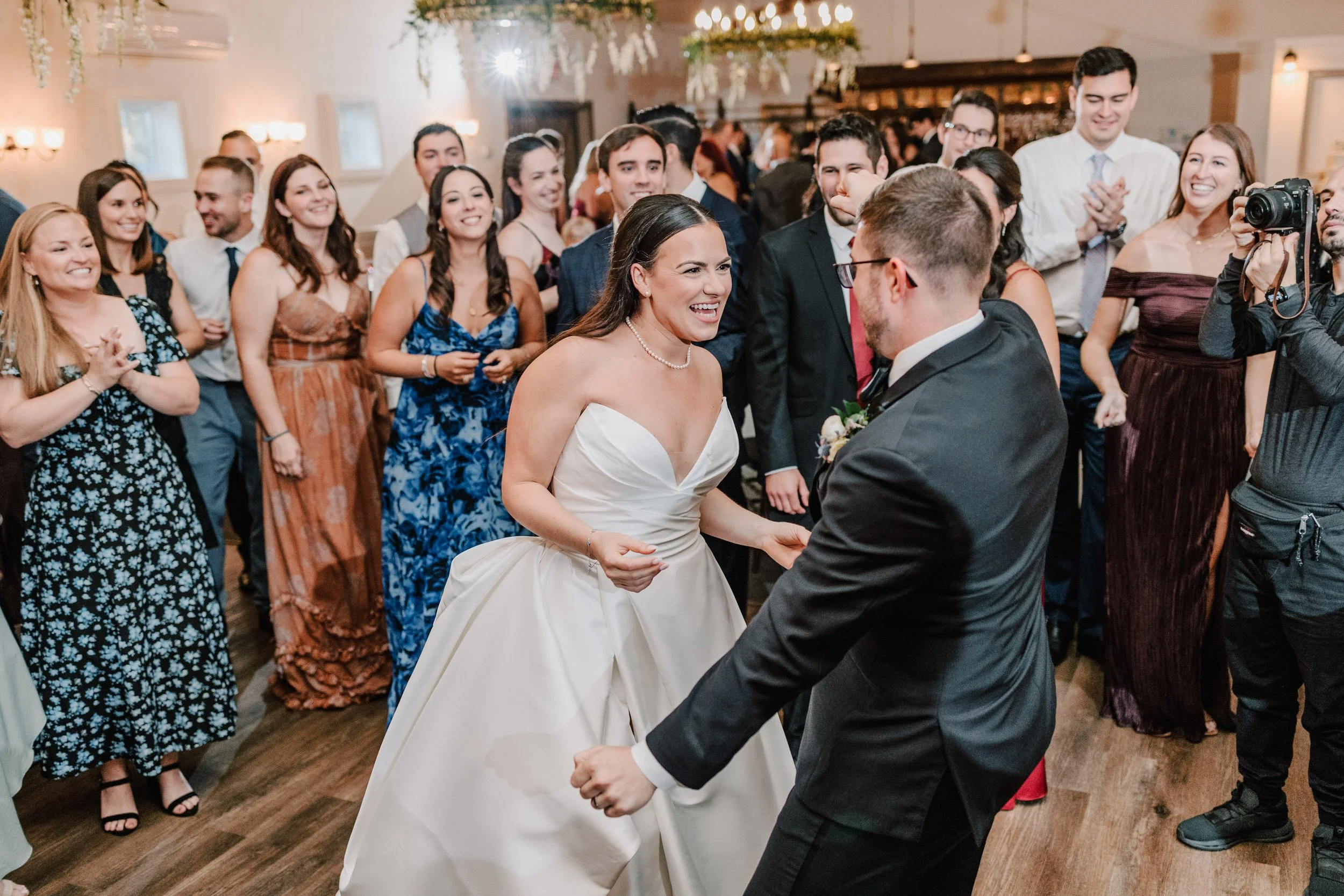 A bride and groom dancing at their wedding reception with guests clapping and smiling in the background.