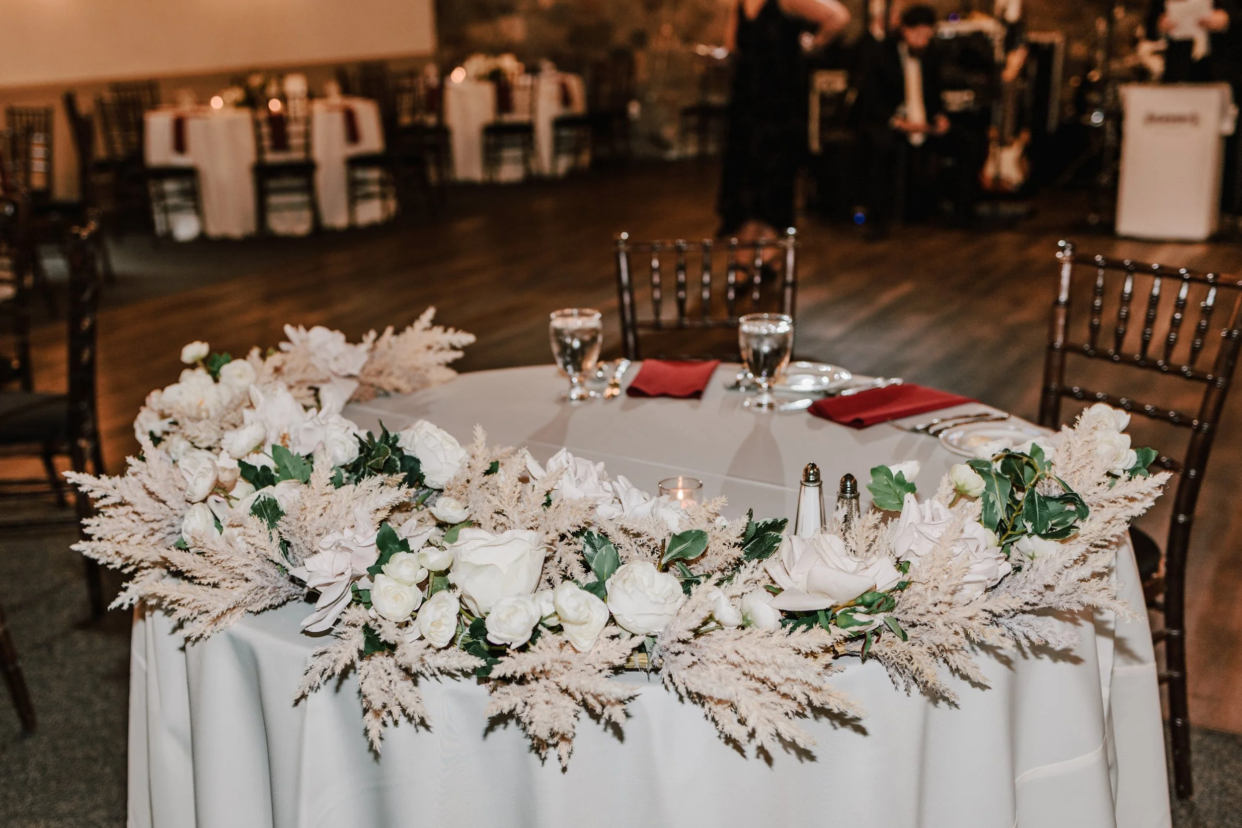Wedding reception table with floral centerpiece, surrounded by chairs with red napkins and glasses of water in dimly lit room.