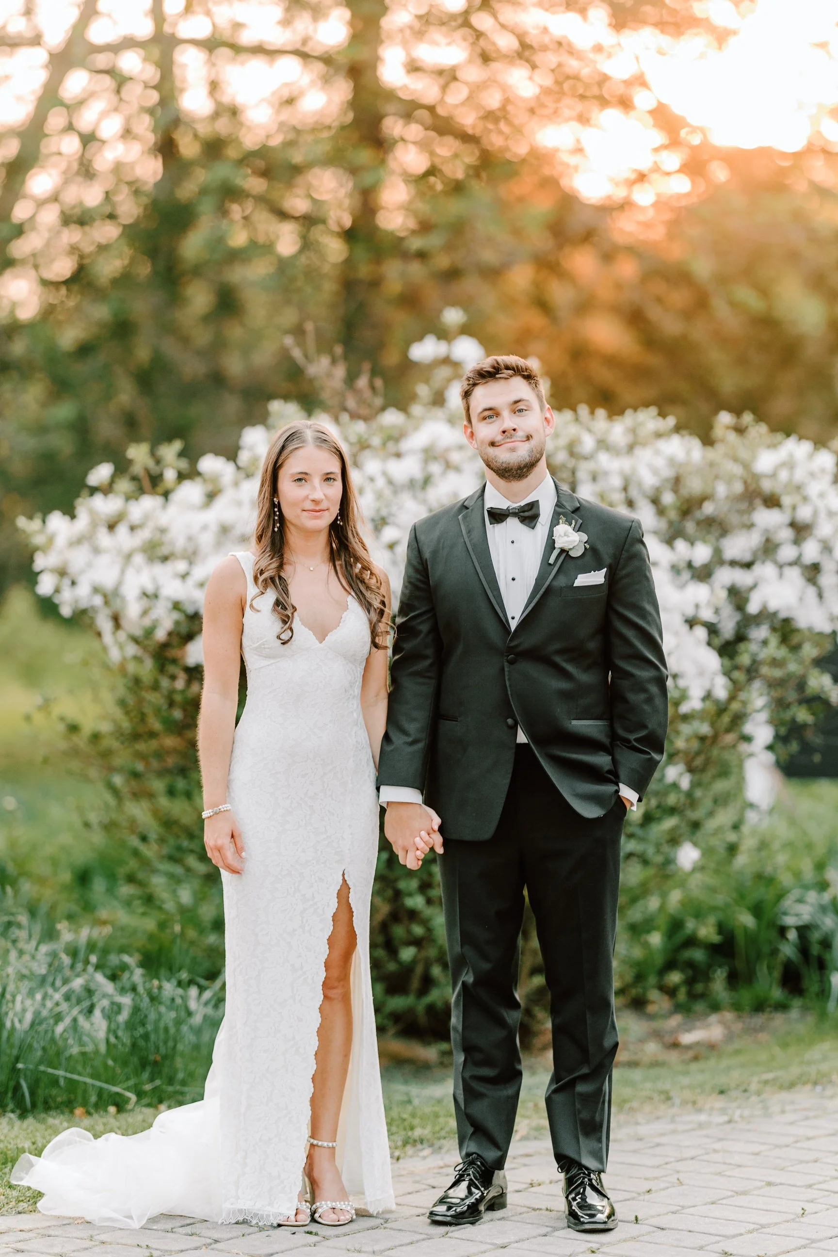A bride and groom stand outdoors holding hands during their wedding. The bride wears a white lace gown with a slit, and the groom wears a black tuxedo with a bow tie. They are in front of a floral backdrop with white flowers, and the sun is setting i