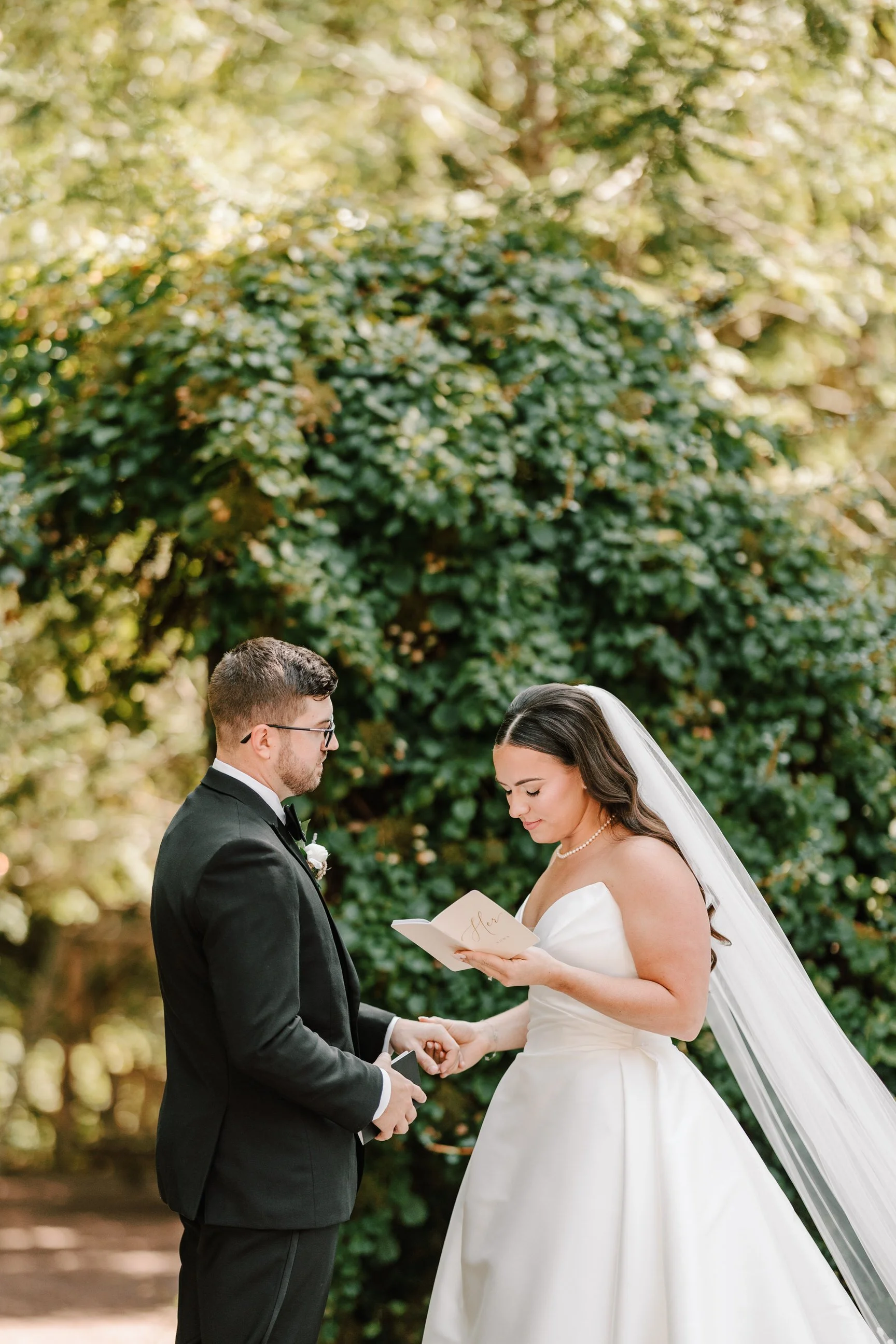 A bride and groom during their wedding ceremony outdoors, holding hands with a green leafy background behind them.