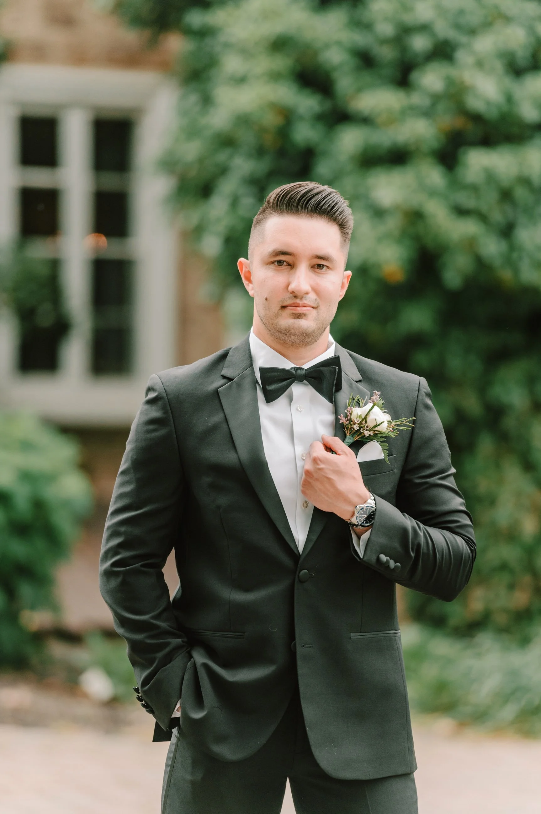 A groom in a black tuxedo with a bowtie and boutonniere stands outdoors in front of green trees and a building, looking at the camera.