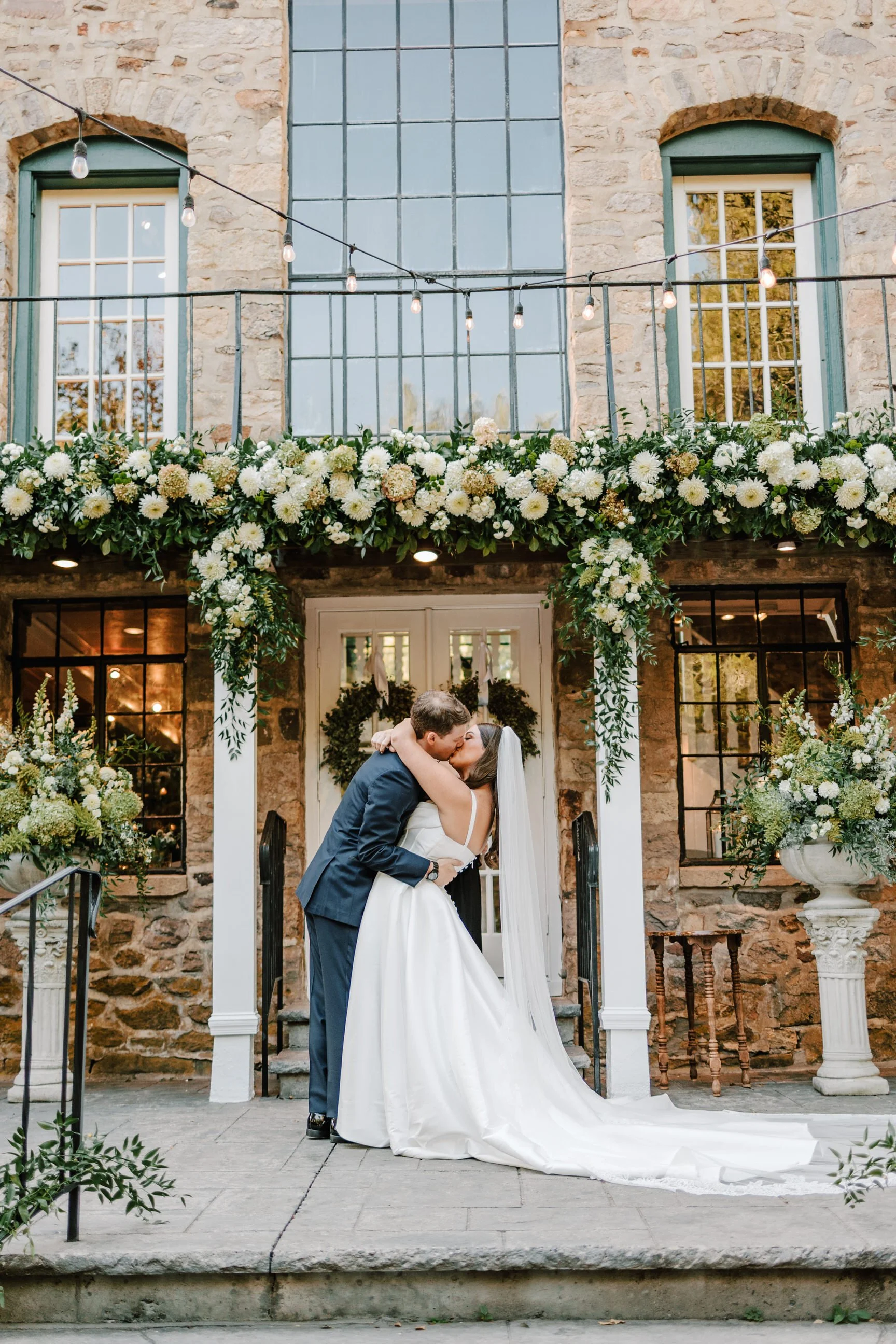 A bride and groom kissing outside of a stone building decorated with white and green flowers, greenery, and string lights for their wedding.