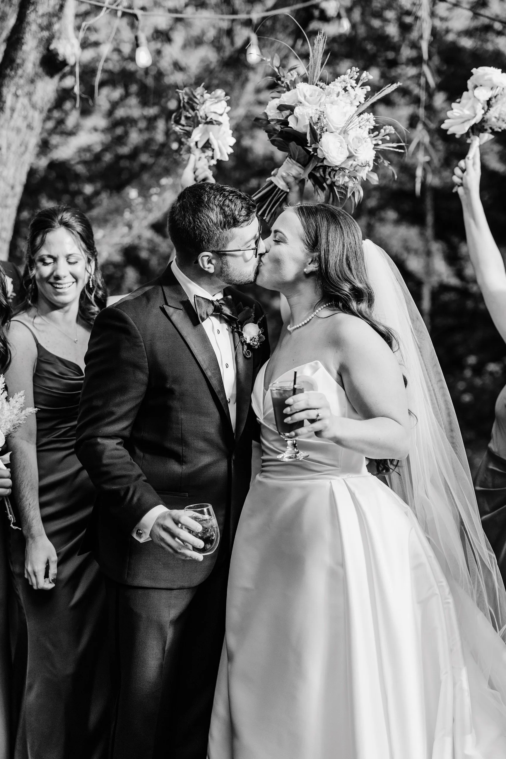Black and white photo of a wedding kiss between a bride in a strapless satin gown and a groom in a tuxedo, surrounded by wedding guests celebrating outdoors.