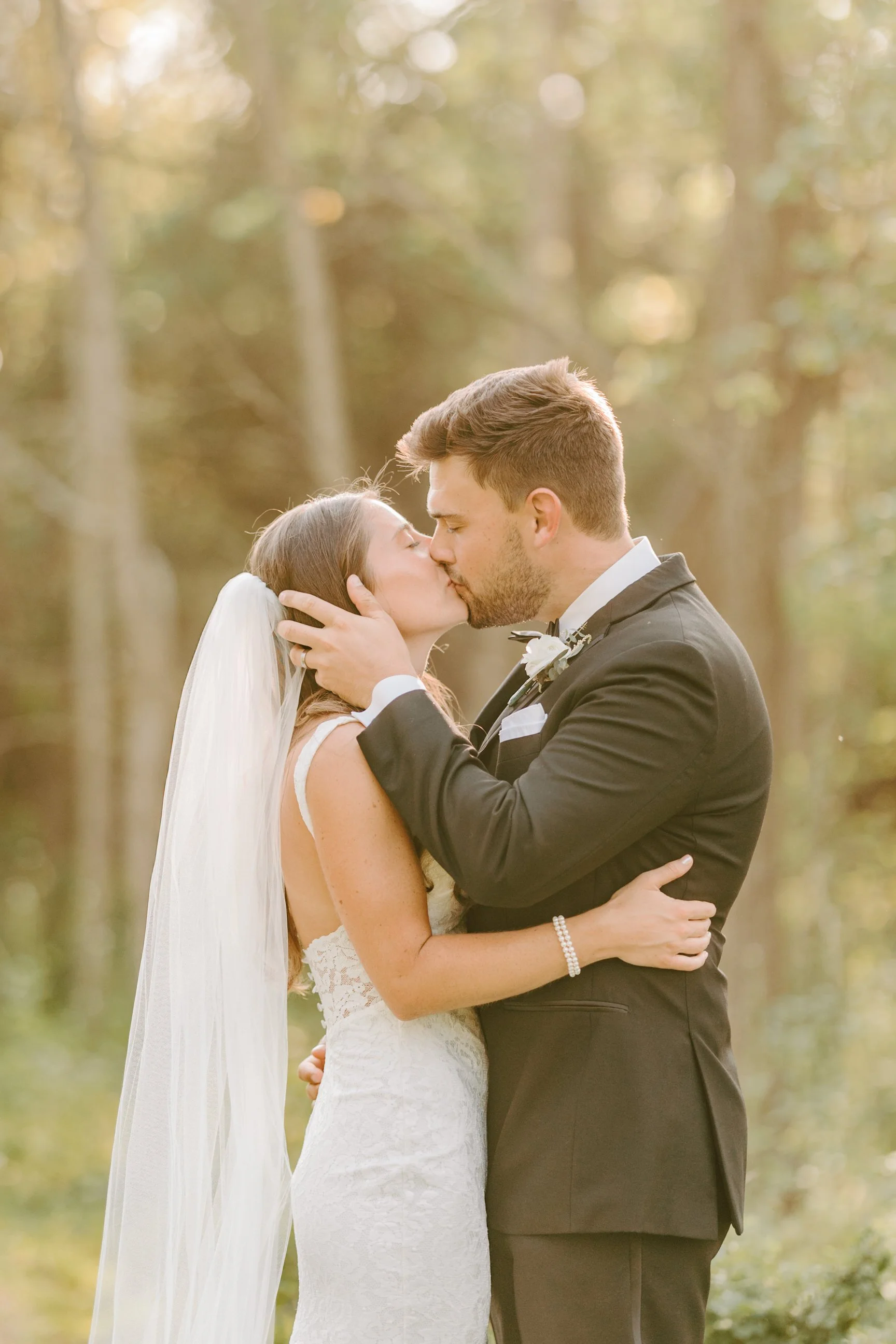 A bride and groom sharing a kiss outdoors, with the bride in a white wedding dress and veil, and the groom in a black tuxedo, during their wedding ceremony or photo session.