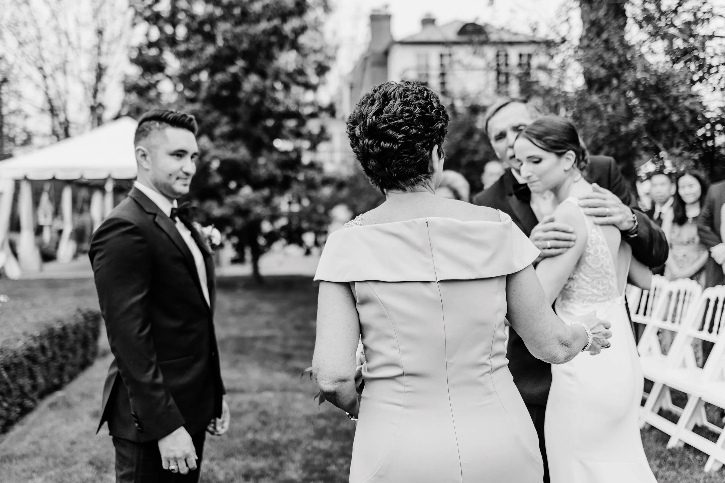 A black and white photo of a wedding scene outdoors. The bride, in a lace dress, is being embraced by a man in a suit, while a woman in a dress stands with her back to the camera and a young man in a tuxedo observes.