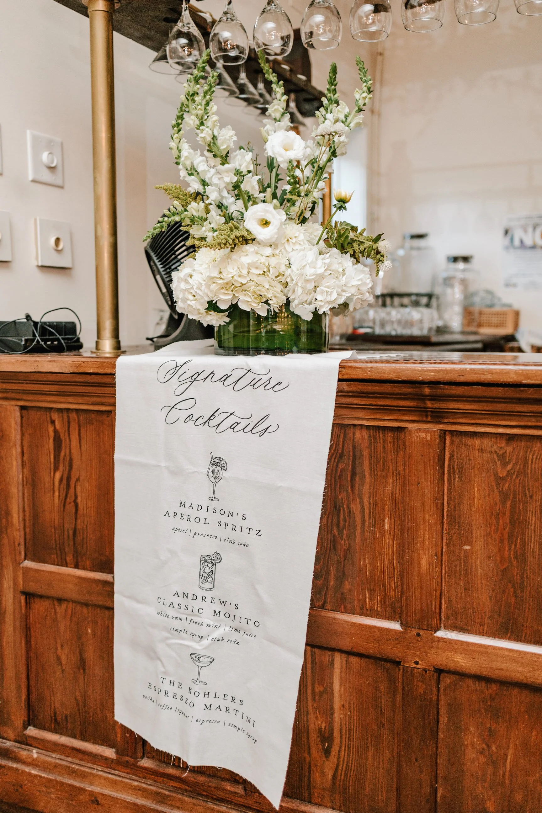 White floral arrangement with white hydrangeas, lisianthus, and greenery on a wooden bar with a white sign listing signature cocktails hanging over it.