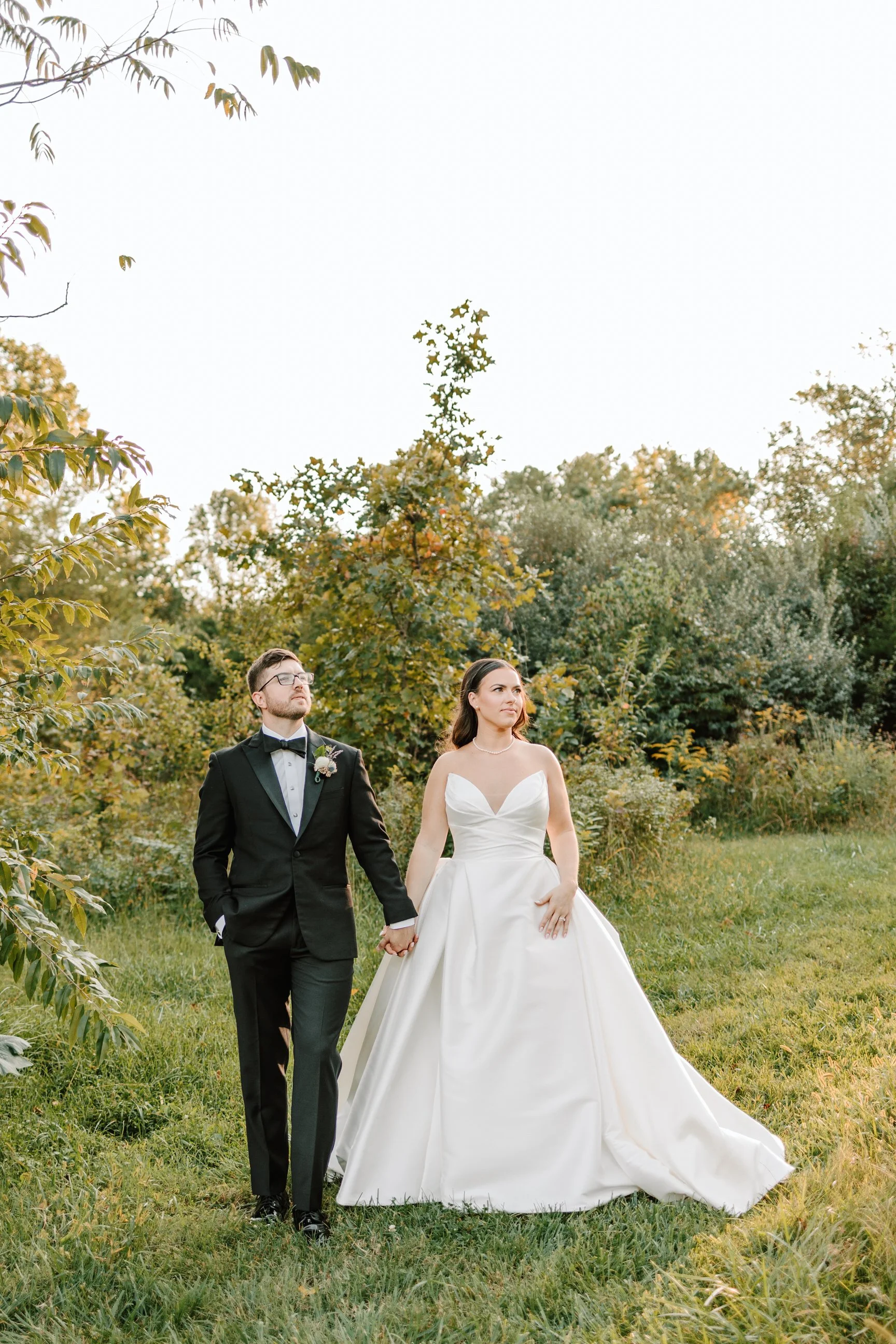 A bride and groom walking hand in hand outdoors, surrounded by green trees and foliage, with the bride wearing a white gown and the groom in a black tuxedo, during a wedding photo shoot.