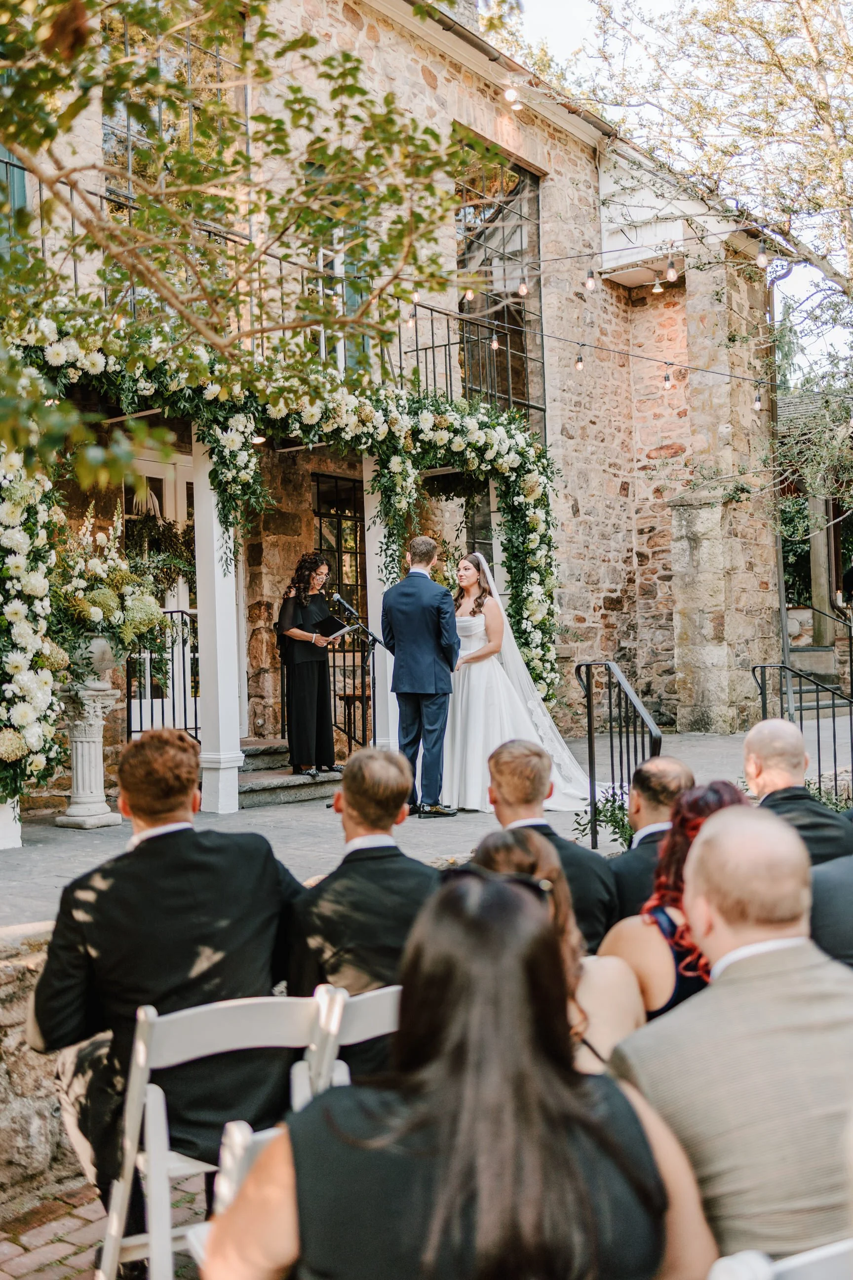 A wedding ceremony taking place outdoors in front of a stone building decorated with white flowers and greenery. The bride and groom are standing before an officiant, exchanging vows, while guests are seated and watching.