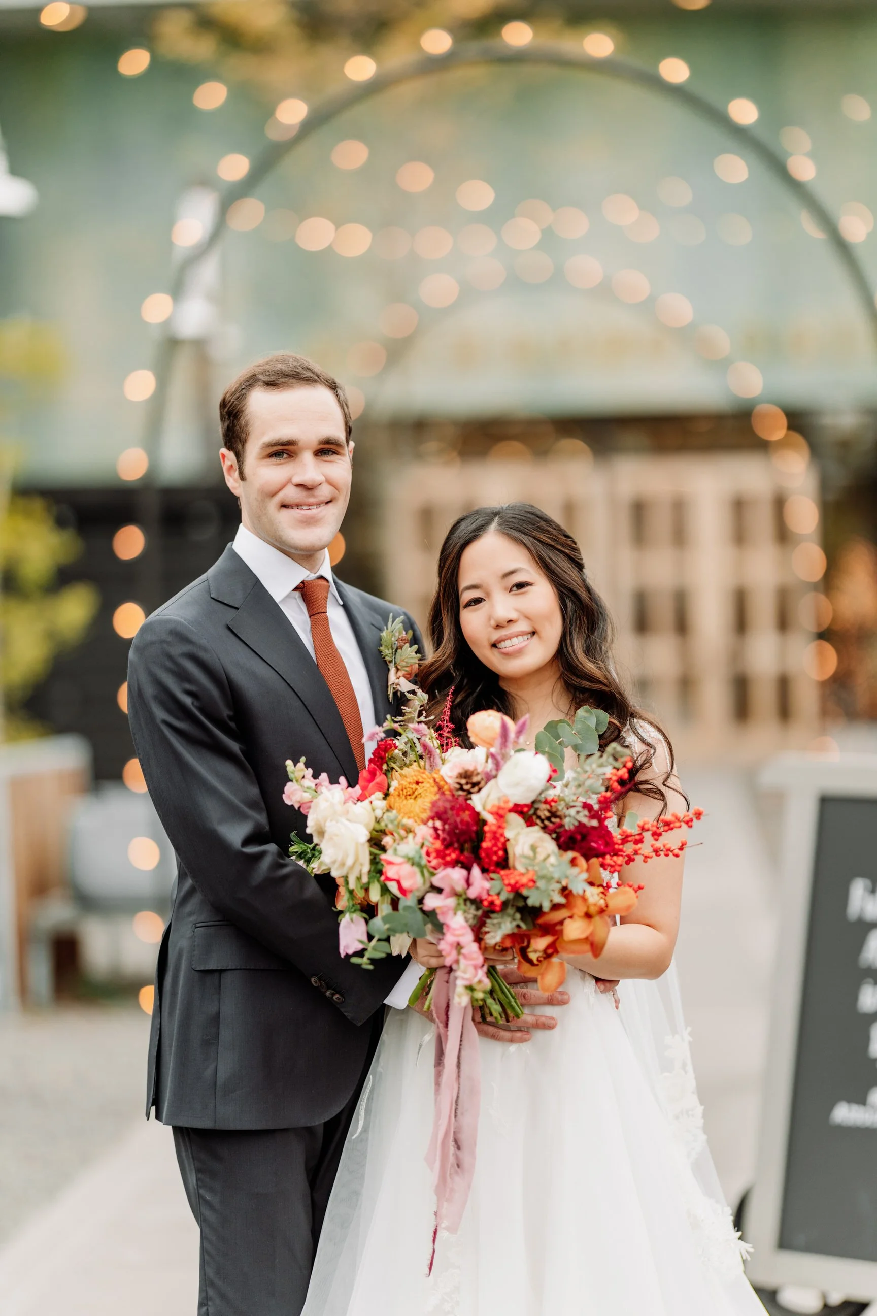 A newlywed couple poses outdoors, with the groom in a gray suit and the bride in a white wedding dress holding a colorful bouquet, under decorative string lights.