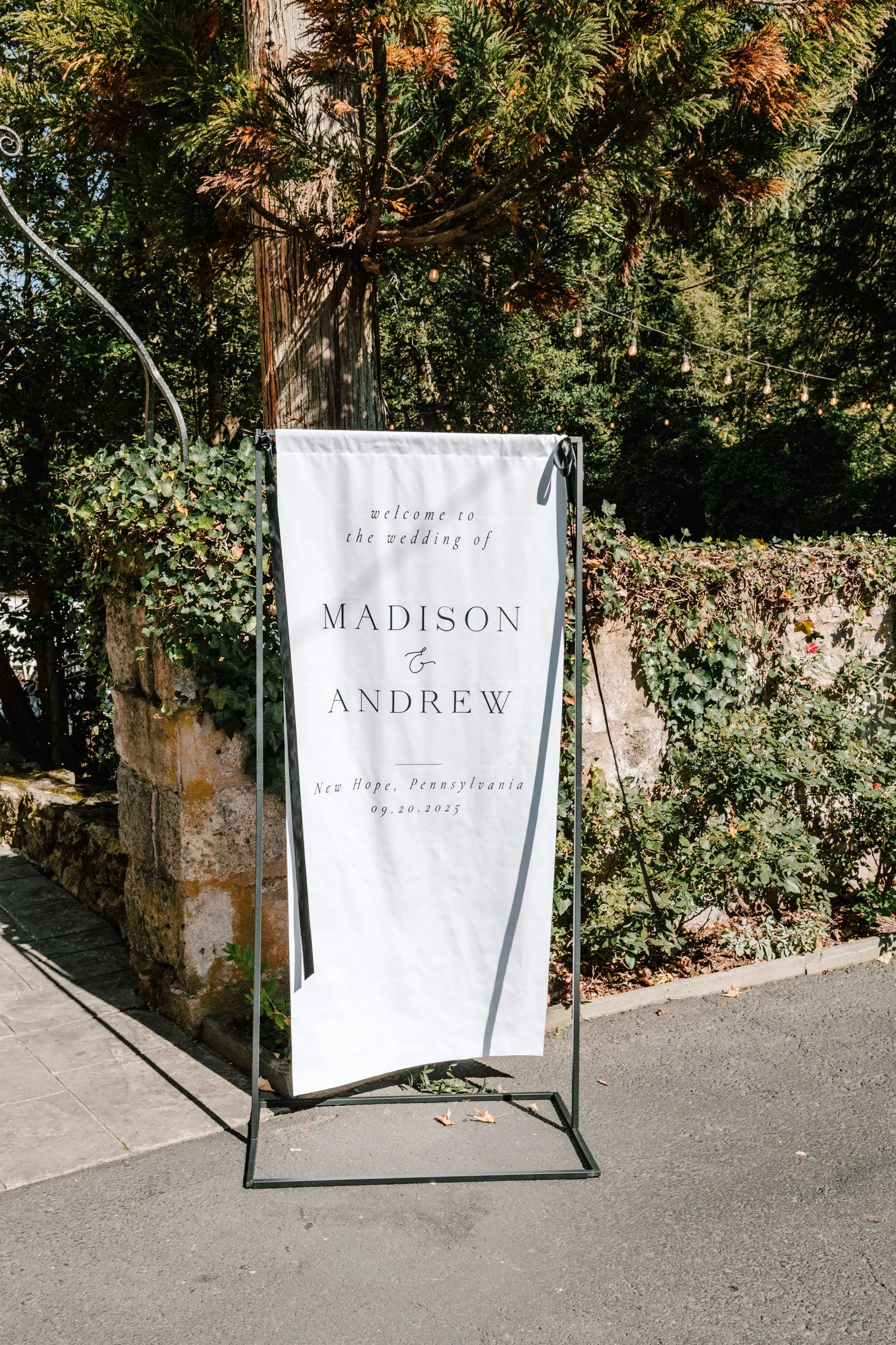 Outdoor wedding sign with black metal frame on gray sidewalk, surrounded by greenery and stone wall, welcoming guests to the wedding of Madison and Andrew in New Hope, Pennsylvania, on September 20, 2025.