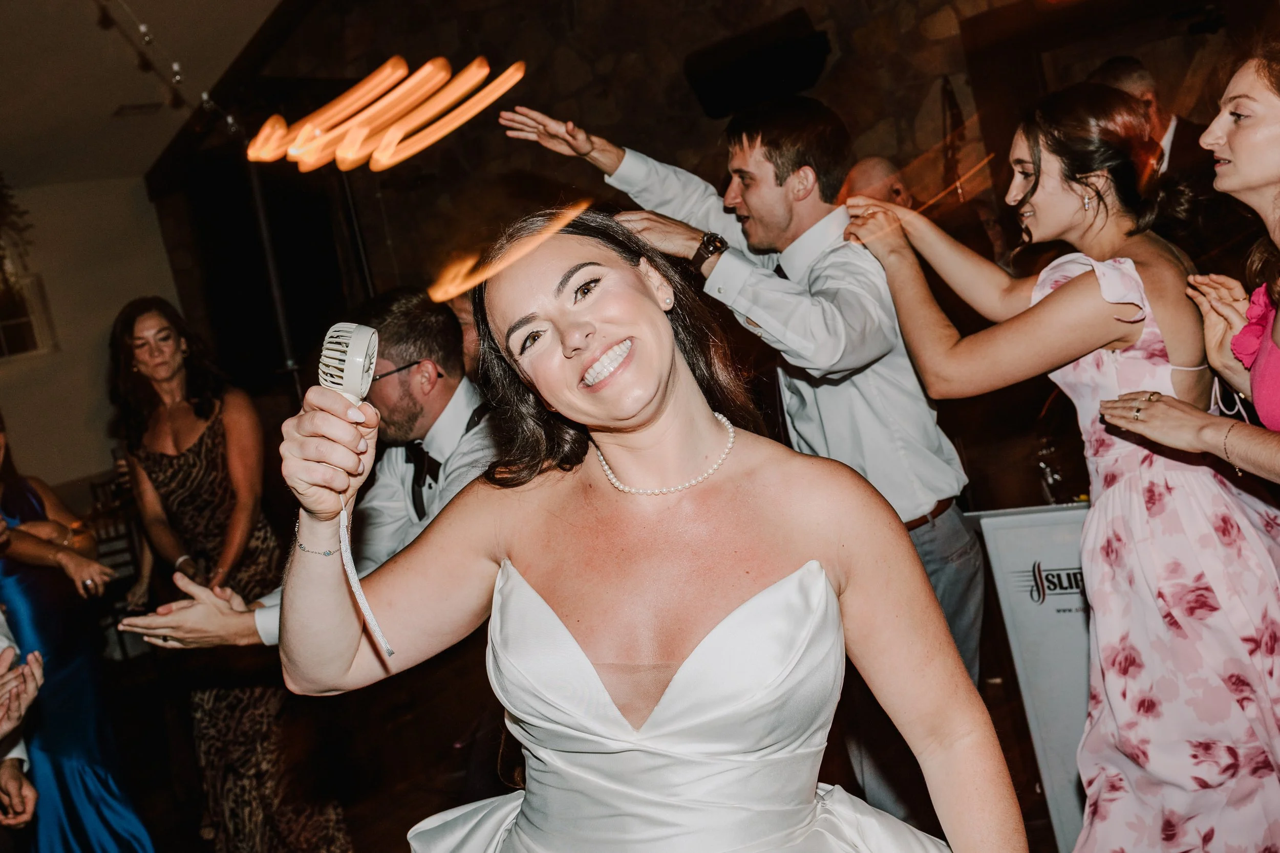 A woman in a wedding dress smiling and holding a microphone at a wedding reception, with guests dancing and celebrating in the background.