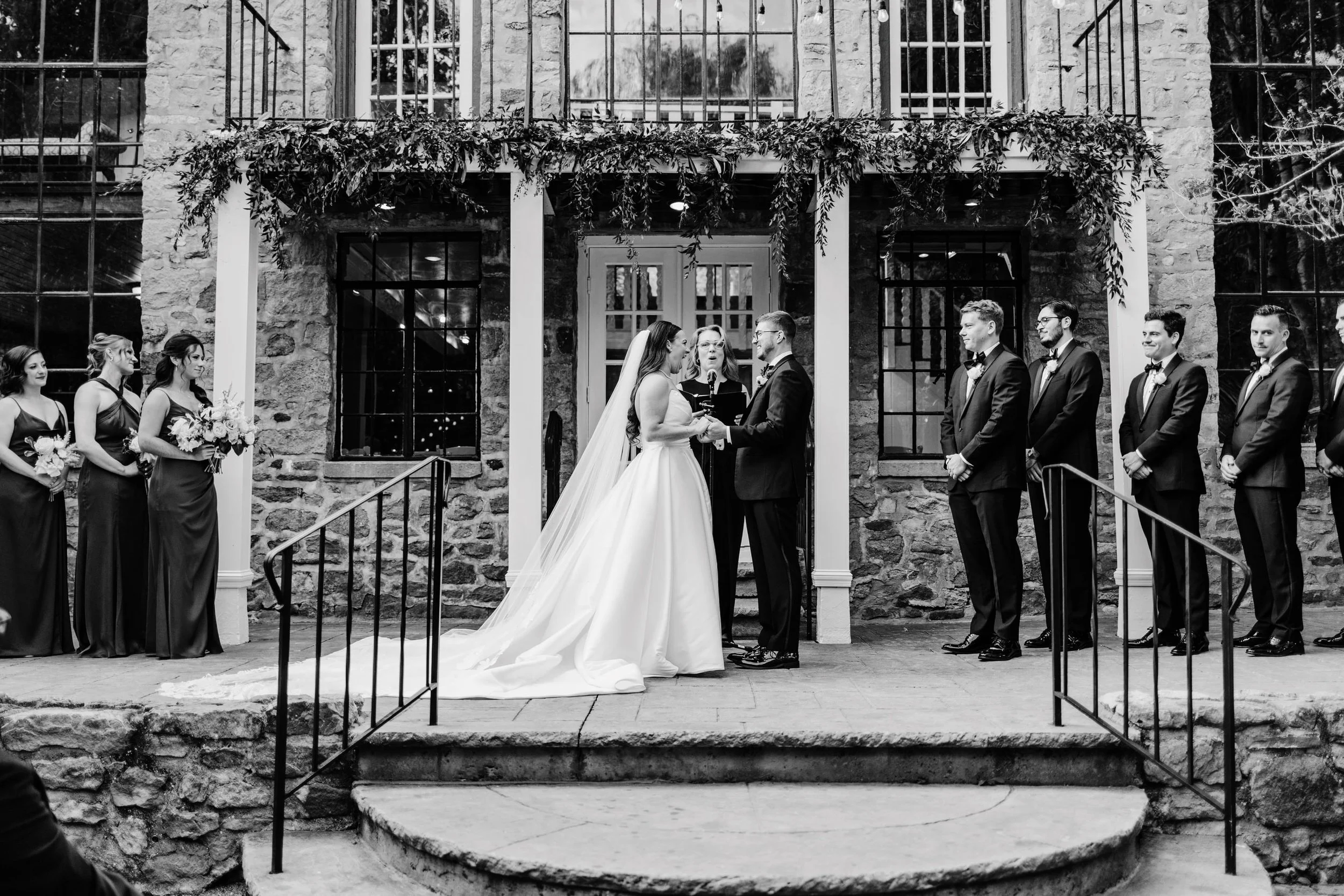 Black and white photo of a wedding ceremony outside a stone building with large windows. The bride and groom are standing in front of a officiant, holding hands and exchanging vows. Bridesmaids and groomsmen are standing on either side, watching the 