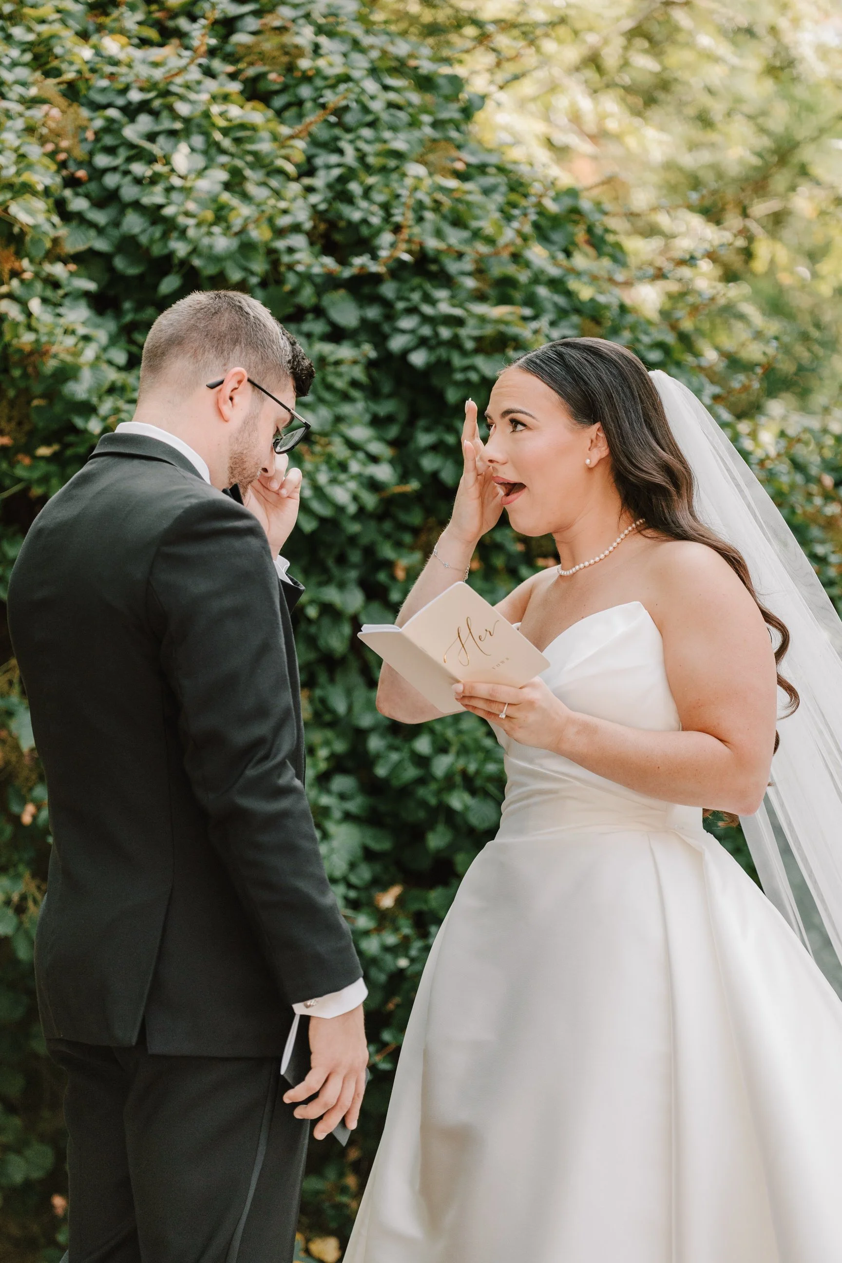 A bride and groom have an emotional moment during their outdoor wedding ceremony. The bride, wearing a white satin wedding gown and pearl necklace, appears to be tearing up as she holds a small book with the word 'Her' on the cover. The groom, dresse