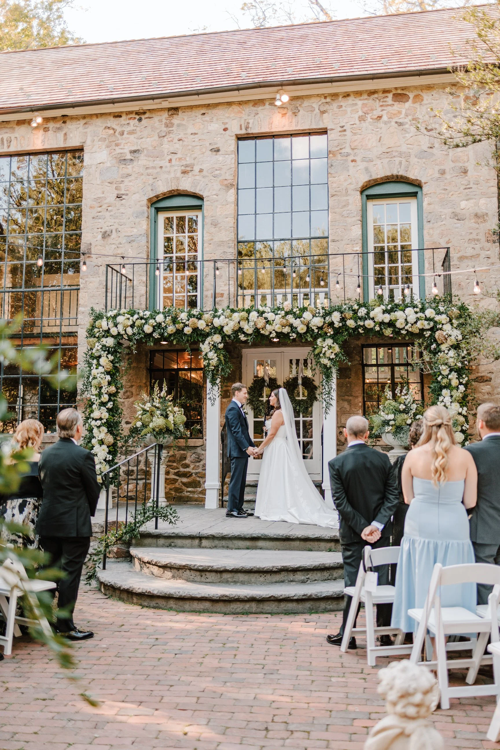Wedding ceremony with bride and groom holding hands in front of a stone building decorated with flowers, with seated guests observing.