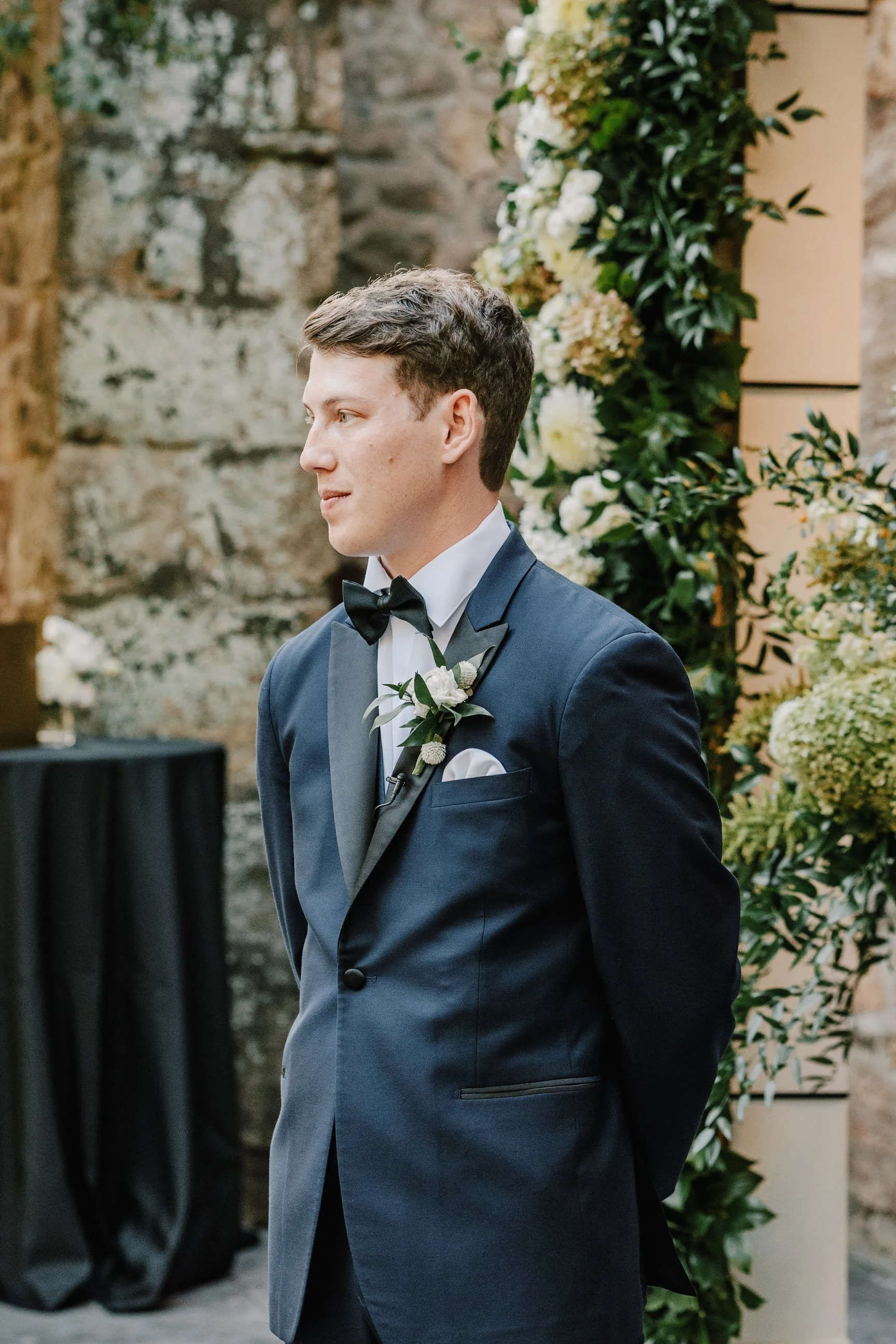 Young man in a tuxedo standing by a floral arrangement during a wedding ceremony.