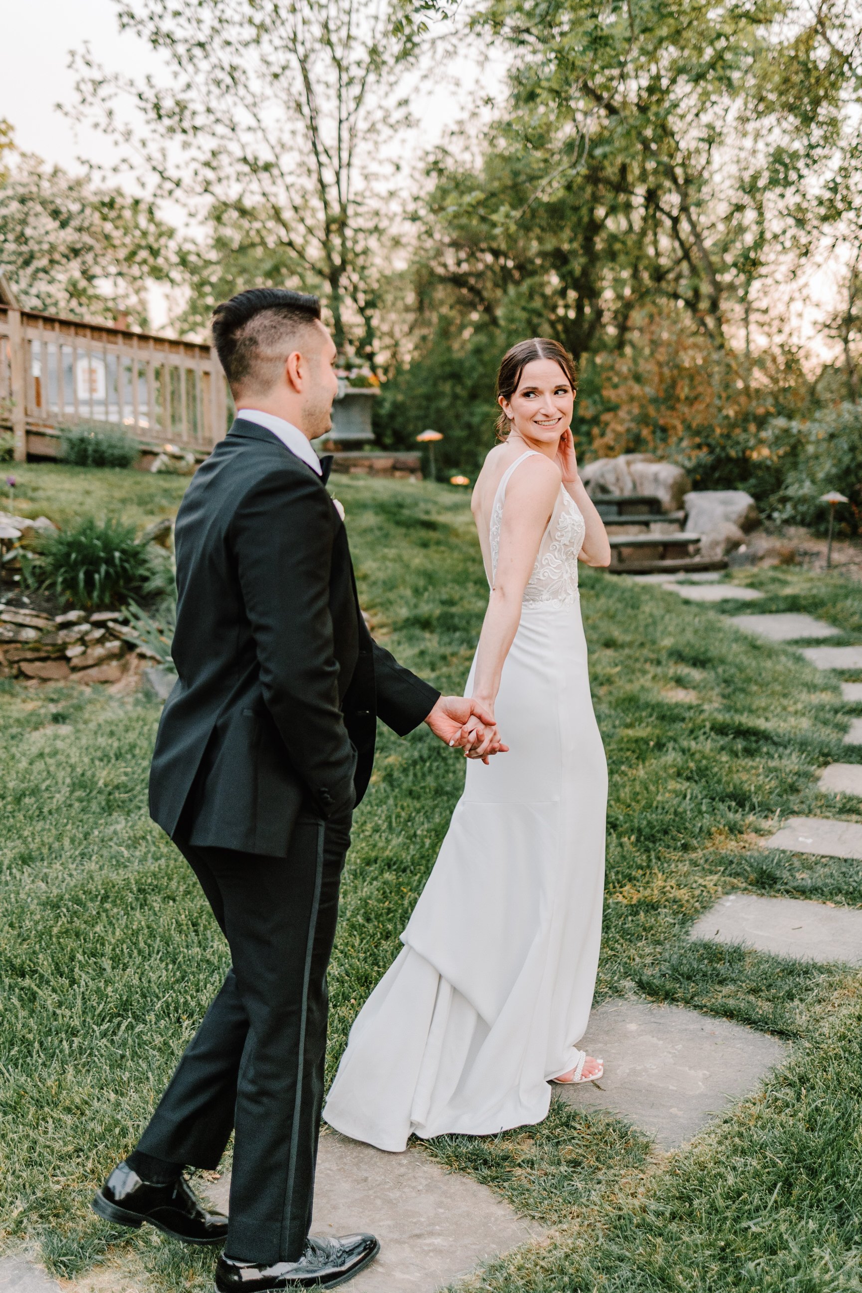 A bride and groom holding hands outdoors on a stone pathway, with trees and greenery in the background during sunset.